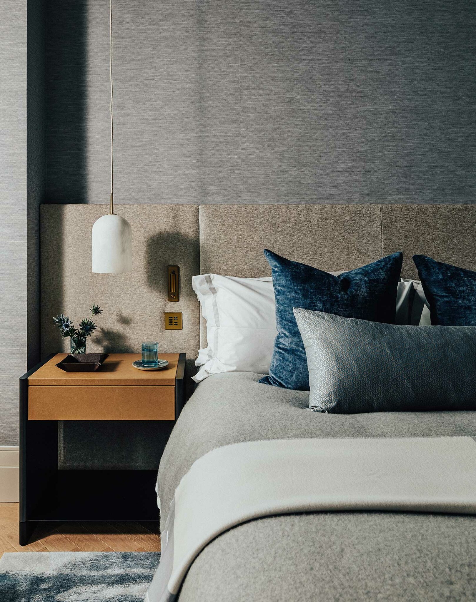 Elegant bedroom with a modern design. Features a grey bed, blue pillows, beige headboard, wooden nightstand, hanging white lamp, creating a calming ambiance.