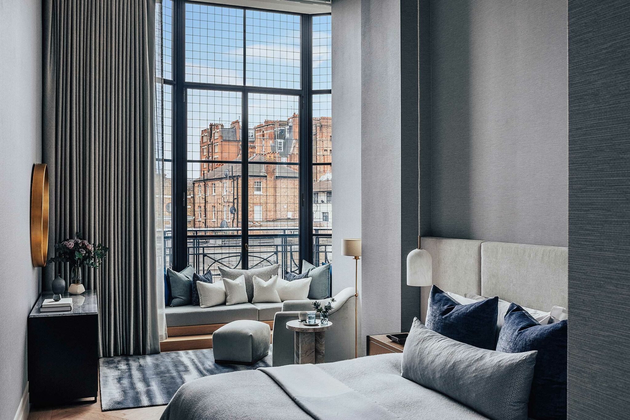 Modern bedroom with large grid windows showing London's cityscape. Neutral tones, plush bed, cozy sofa with cushions, and a soft ambiance.