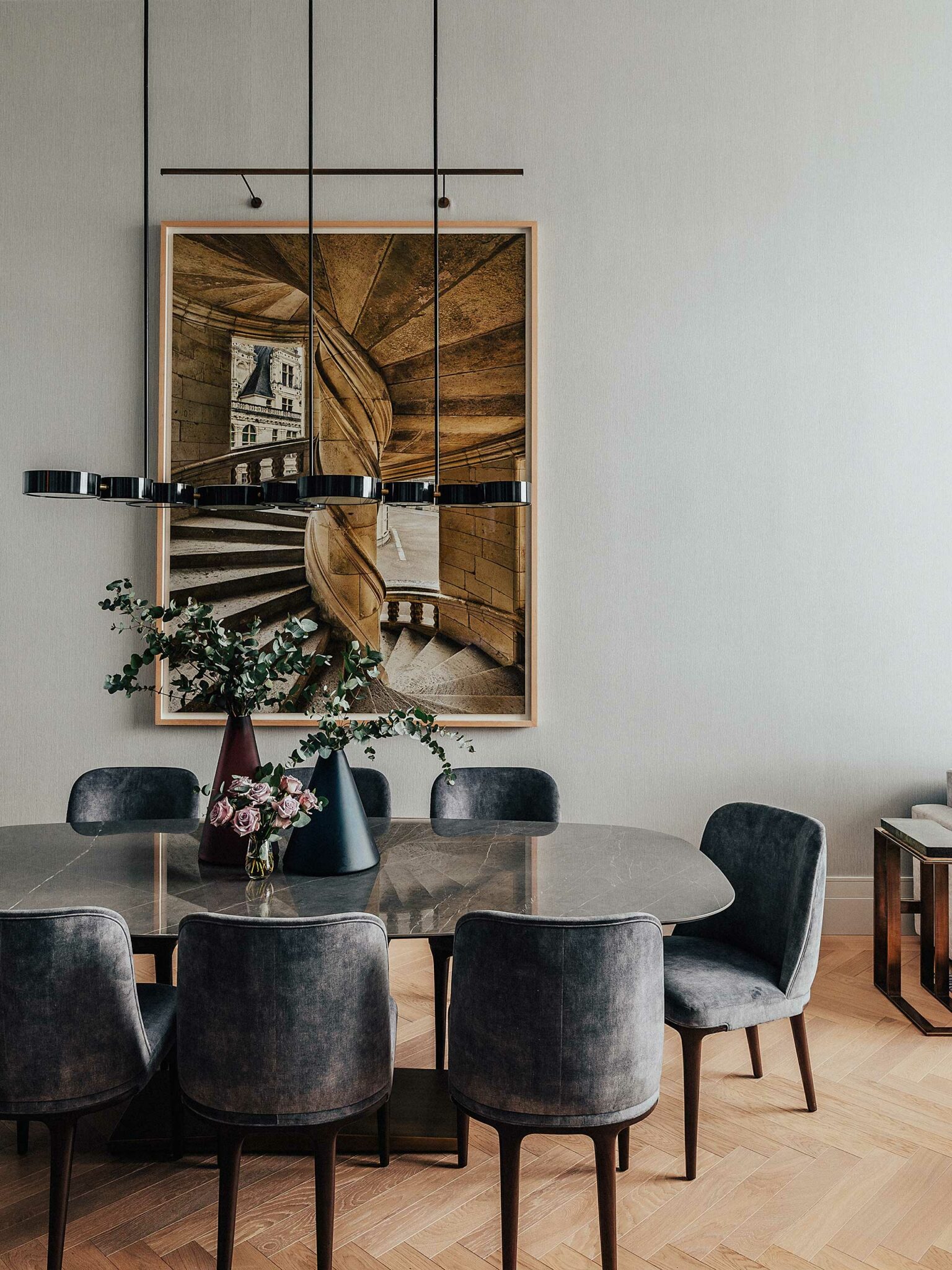 A modern dining room with a gray oval table, eight upholstered chairs, and two vases of flowers. A large spiral staircase painting and sleek pendant light add elegance.