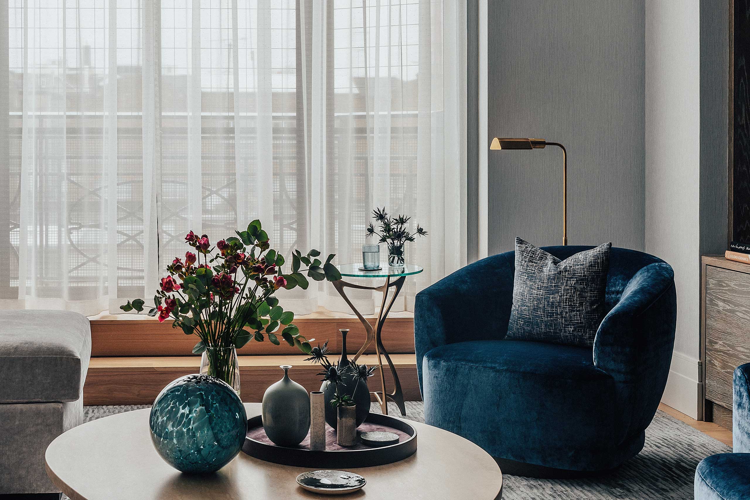 Cozy living room with a blue velvet chair, gray pillow, and a round table with vases and flowers. Tall windows with sheer curtains in the background.