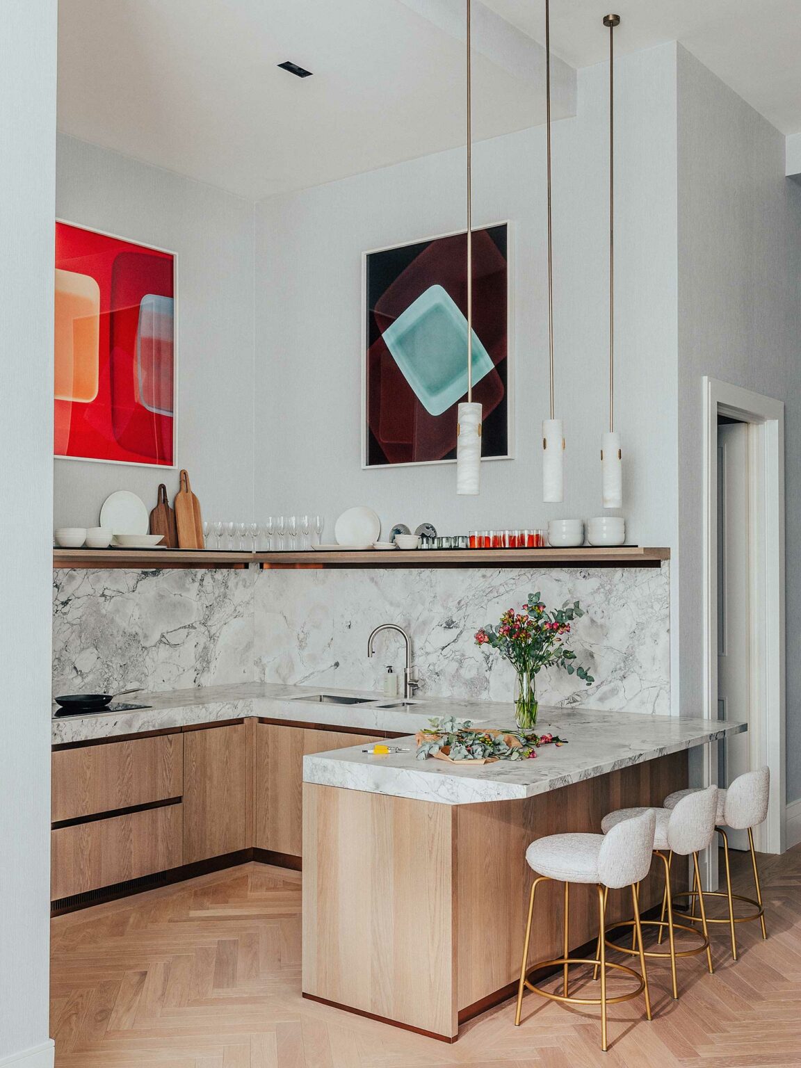 Modern kitchen with wooden cabinets, marble countertops, and three pendant lights. Abstract red and blue paintings adorn the walls, creating a chic and vibrant atmosphere.