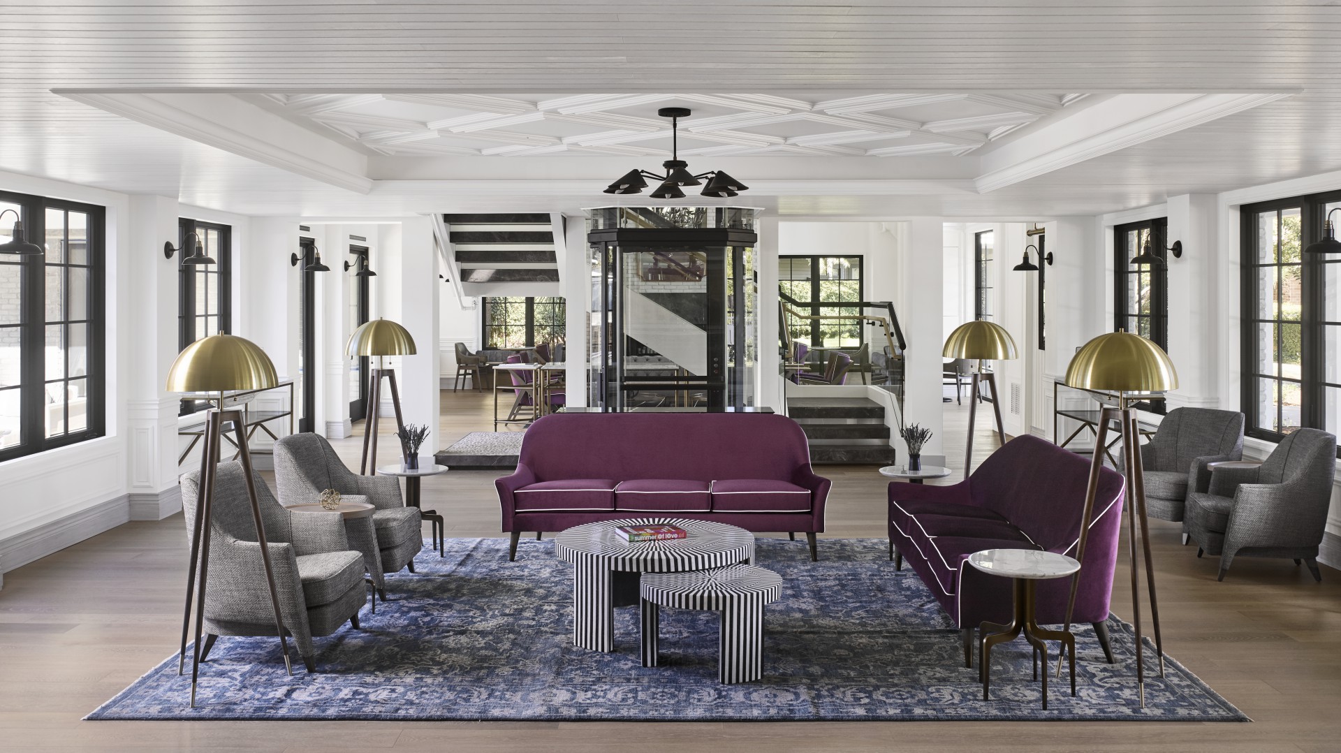 A modern hotel lounge with purple sofas, gray armchairs, and a blue patterned rug. Gold floor lamps and striped tables add elegance. Bright, spacious ambiance.