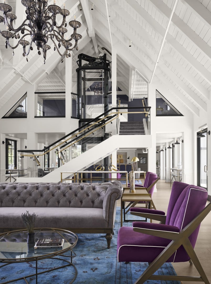 Spacious modern hotel lounge with high vaulted ceiling, a glass elevator, and elegant black chandelier. Features gray sofa, purple chairs, and blue rug.