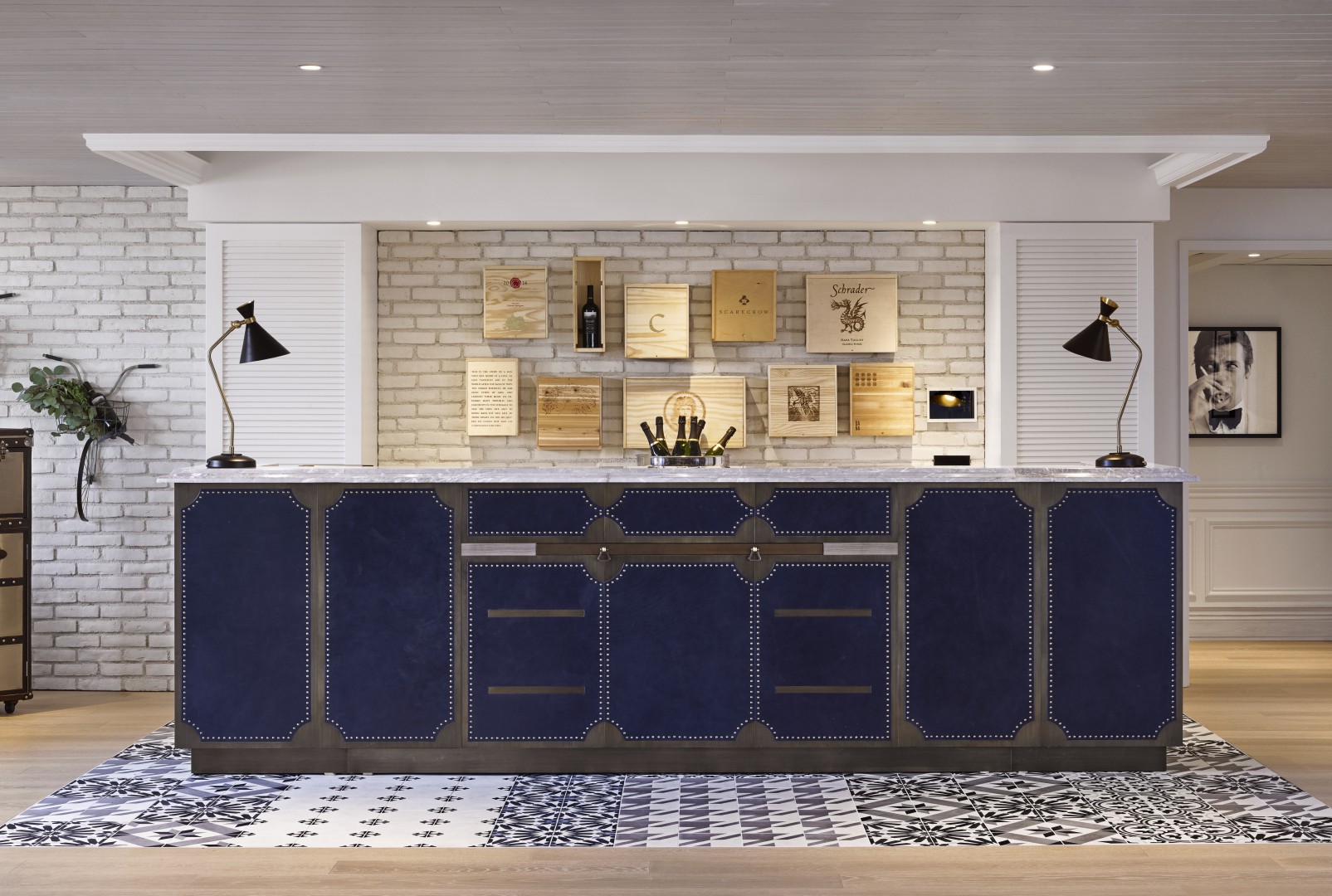 Elegant hotel reception with a navy blue cabinet featuring silver accents, a patterned black-and-white tiled floor, and a white brick wall adorned with wooden plaques.