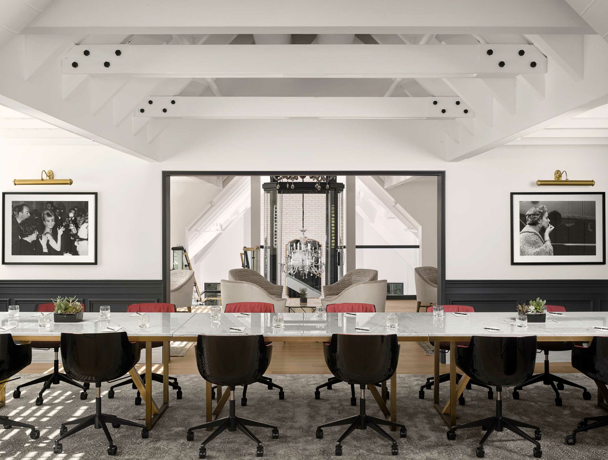 A sleek hotel conference room with a long table, black chairs, and red accents. Black-and-white photos adorn the walls. A cozy lounge with armchairs is visible through a doorway.