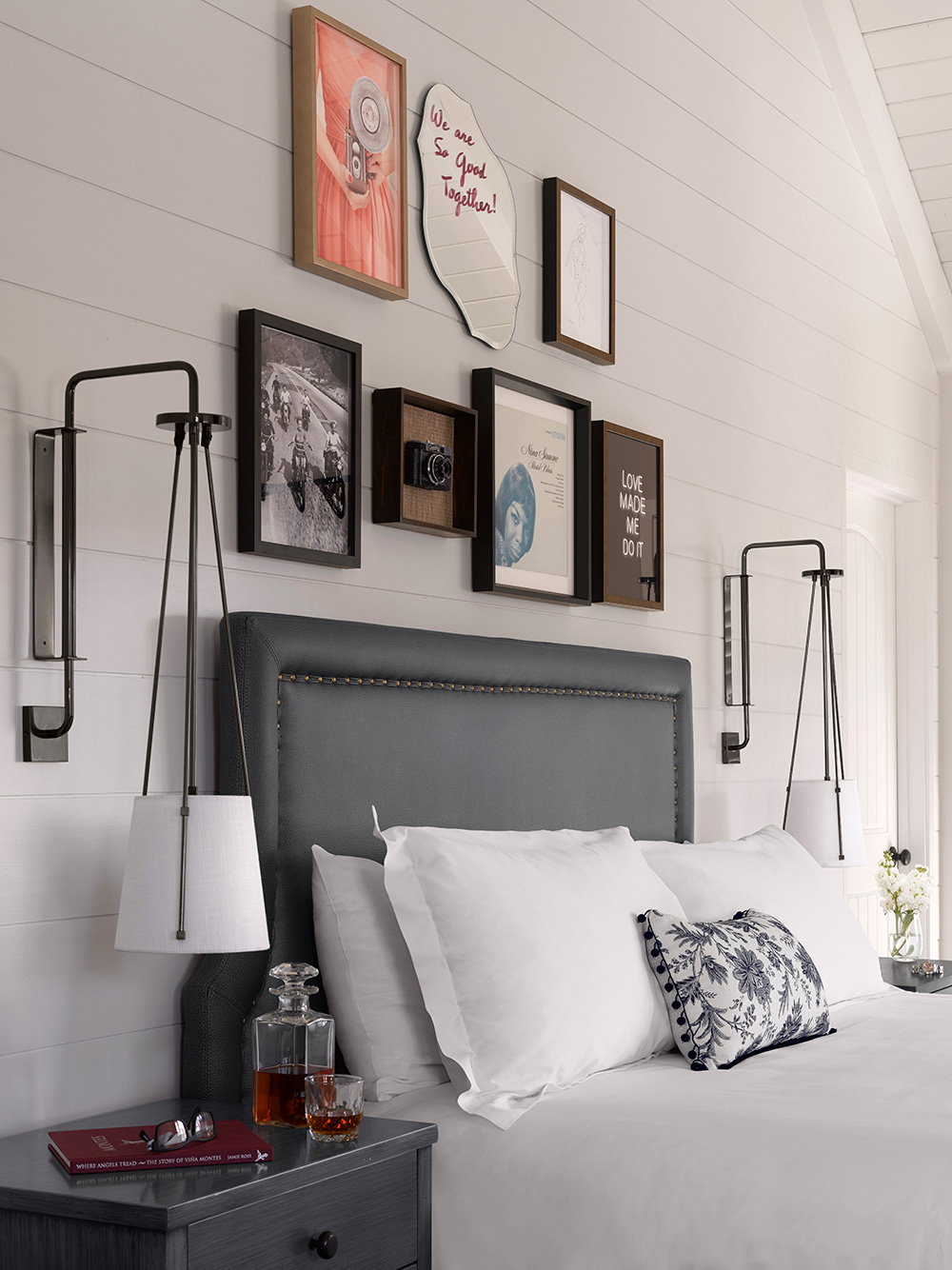 A cozy hotel bedroom features a bed with white pillows and a floral pillow. Above, a gallery wall displays vintage photos, art, and quotes. The space feels warm and inviting.