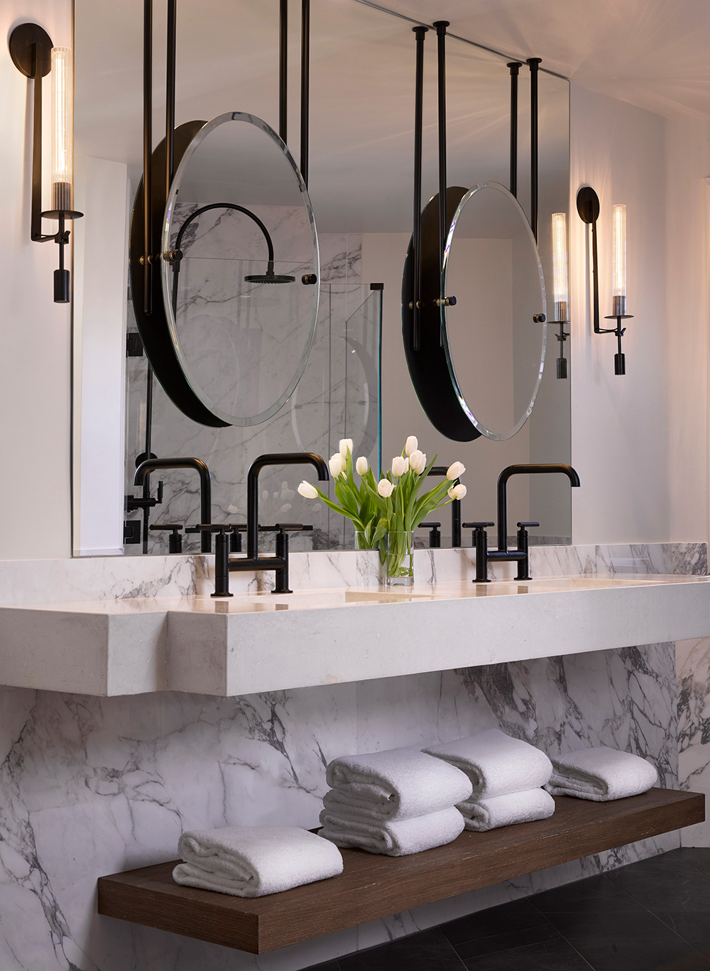 Modern suite bathroom with dual black faucets and round mirrors on a marble vanity. Fresh white tulips in a vase add elegance. Neatly folded towels below.
