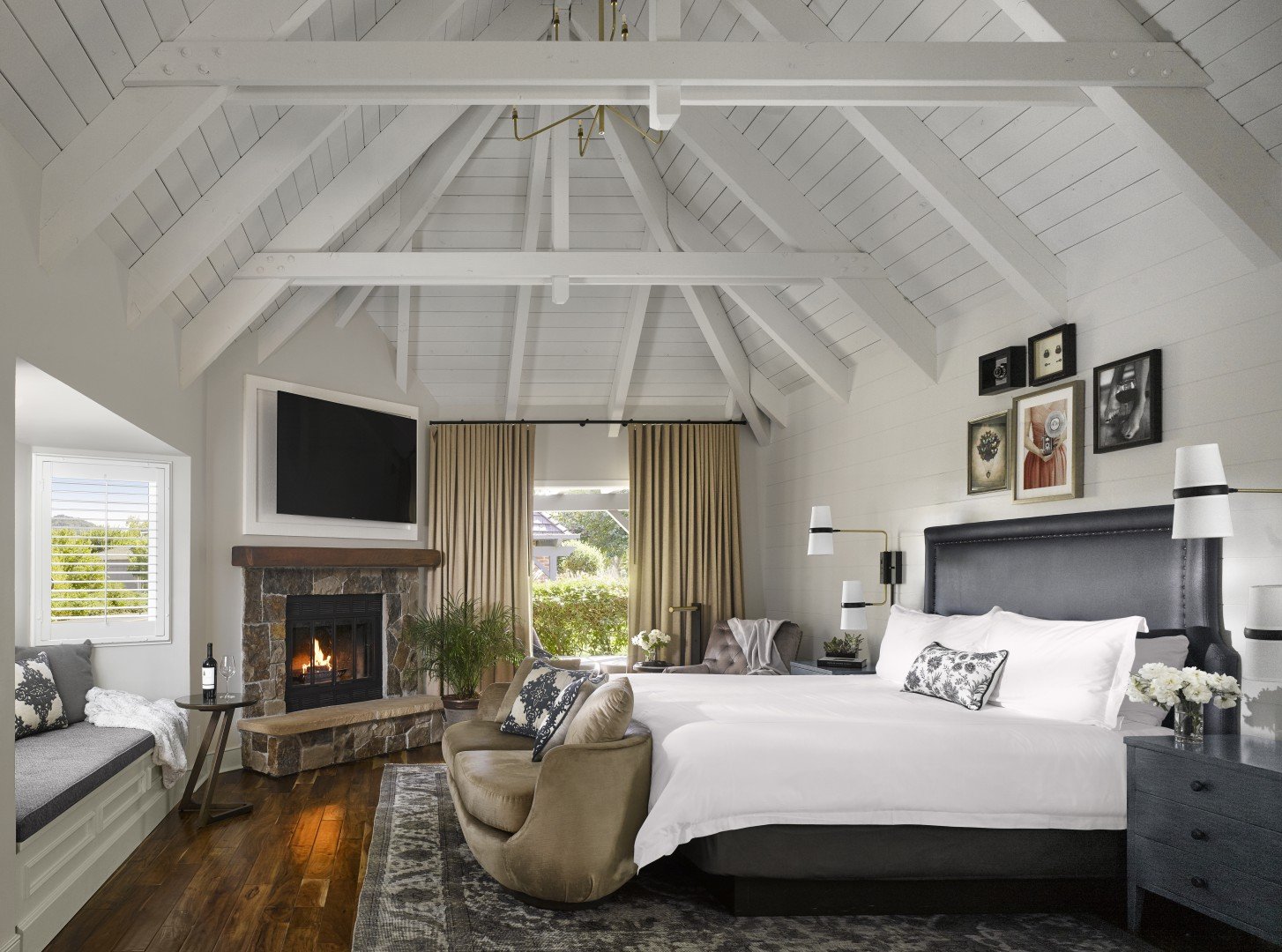 Cozy hotel suite with vaulted ceiling, fireplace, and window seat. Neutral tones, framed art, and elegant furnishings create a serene, inviting atmosphere.