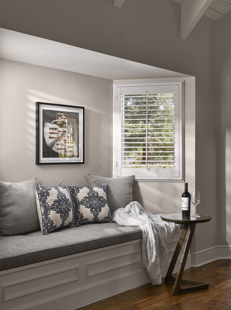 A cozy suite nook with a gray cushioned bench, blue patterned pillows, and a soft throw. A small table holds a wine bottle and glass. A window offers natural light.