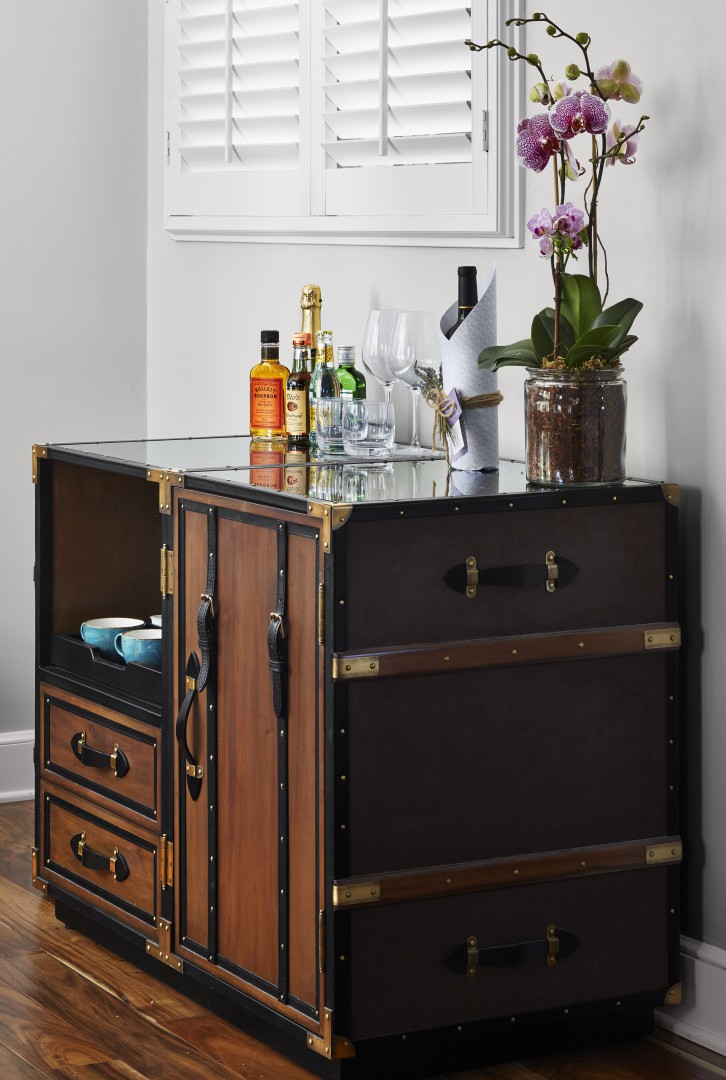Vintage-style bar cart with wooden panels, glass top holding assorted liquor bottles, wine glasses, a linen napkin, and a potted orchid. Bright, tidy ambience.