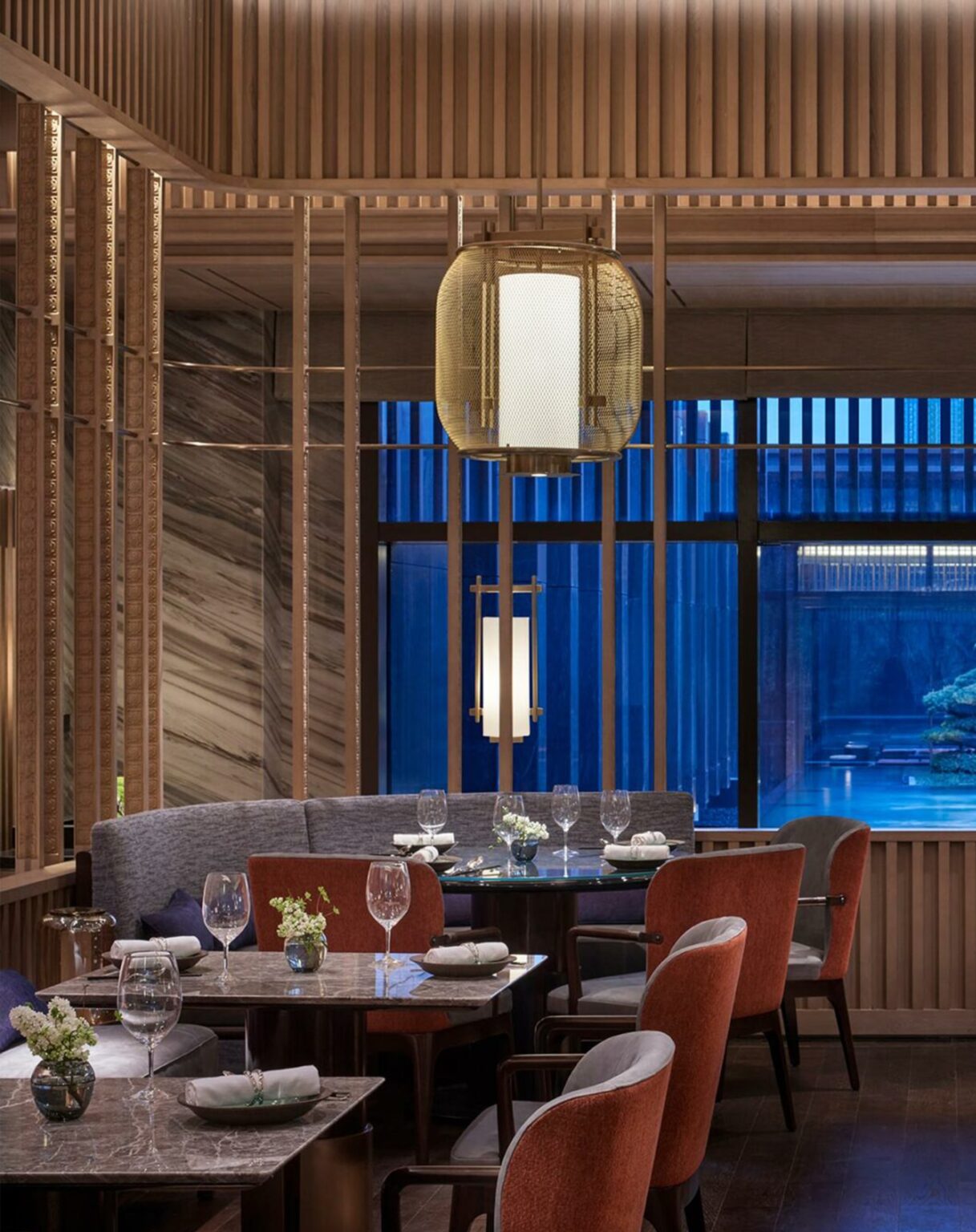 Sophisticated hotel restaurant interior with elegant wooden panels, red and gray upholstered chairs, soft lighting, and a serene view of a Japanese garden.