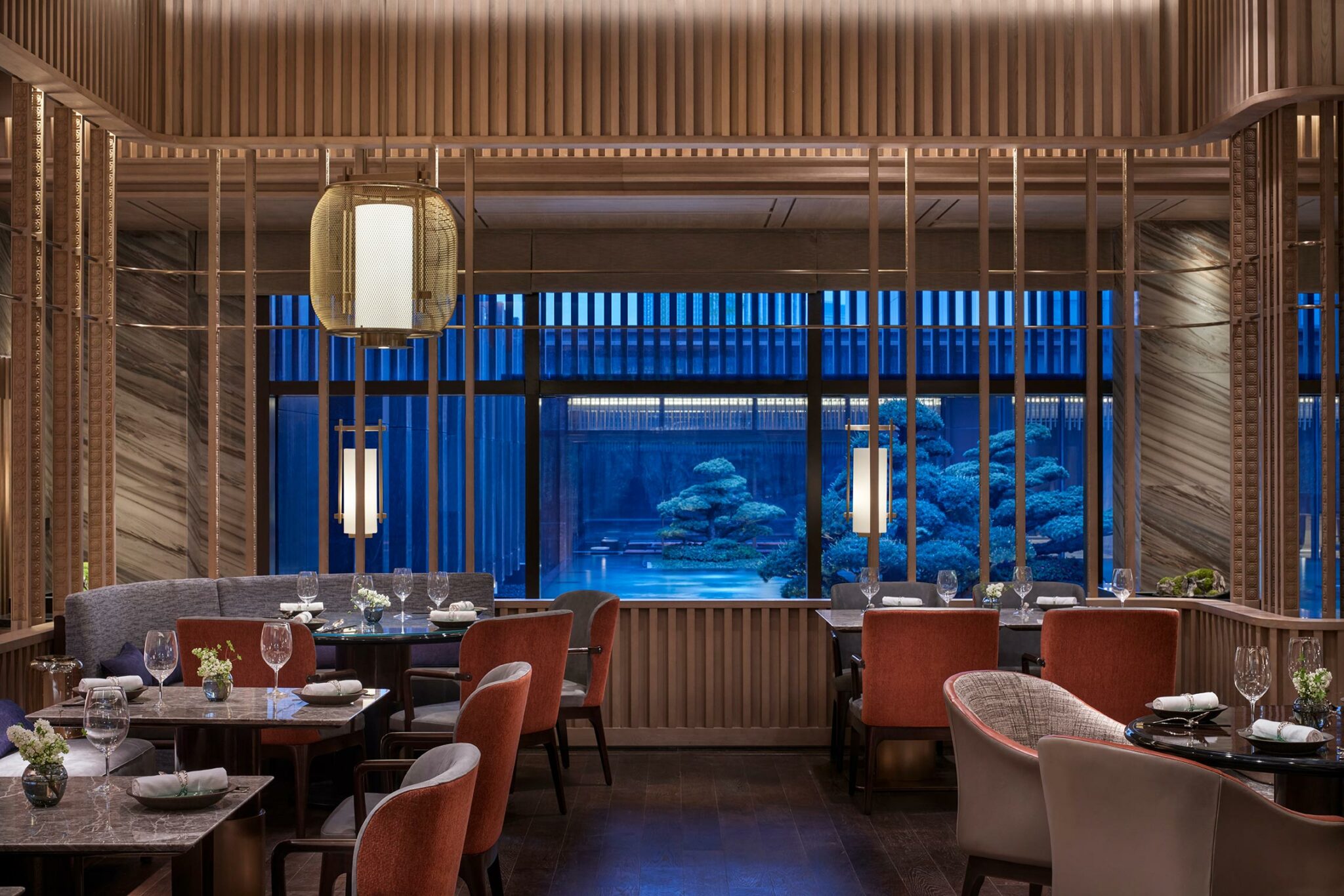 Sophisticated hotel restaurant interior with elegant wooden panels, red and gray upholstered chairs, soft lighting, and a serene view of a Japanese garden.