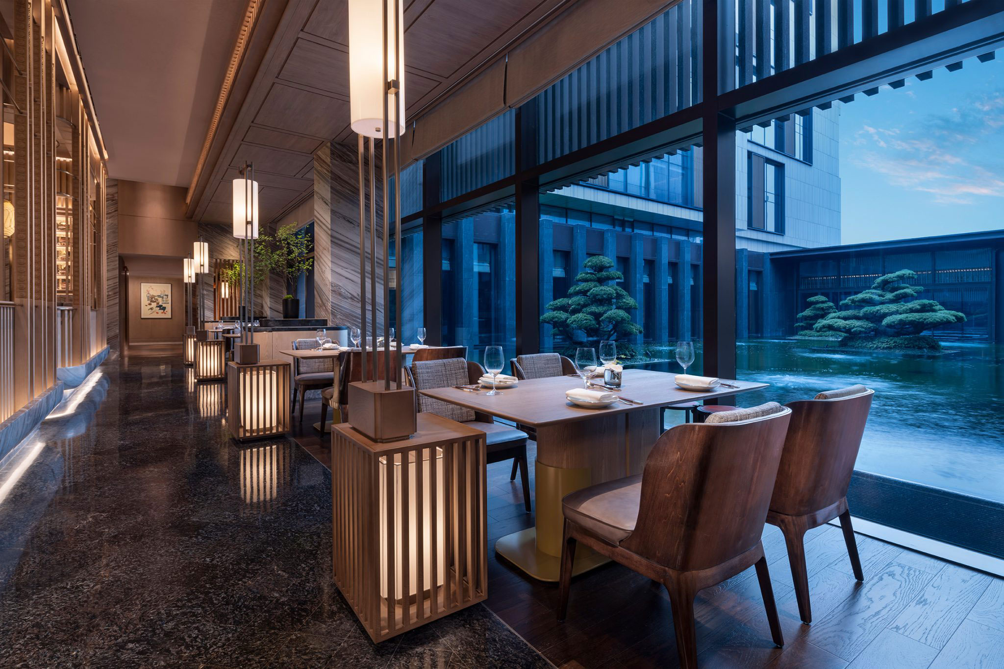 Elegant hotel restaurant with wooden decor, soft lighting, and large windows overlooking a serene water garden. Atmosphere is calm and luxurious.
