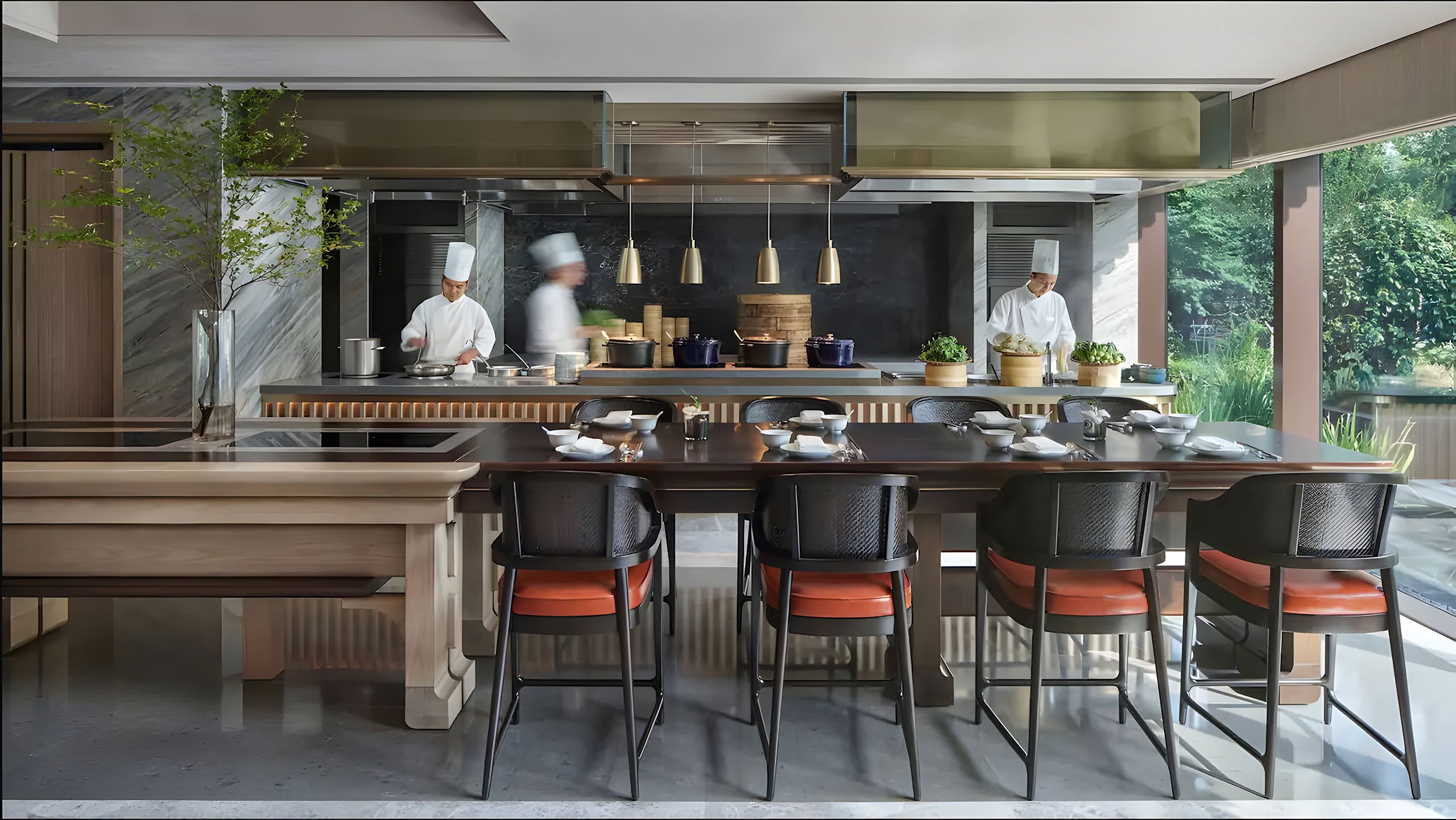 Modern hotel restaurant kitchen with two chefs in white uniforms working. A long table with black chairs is set with dishes. The atmosphere is elegant and busy.