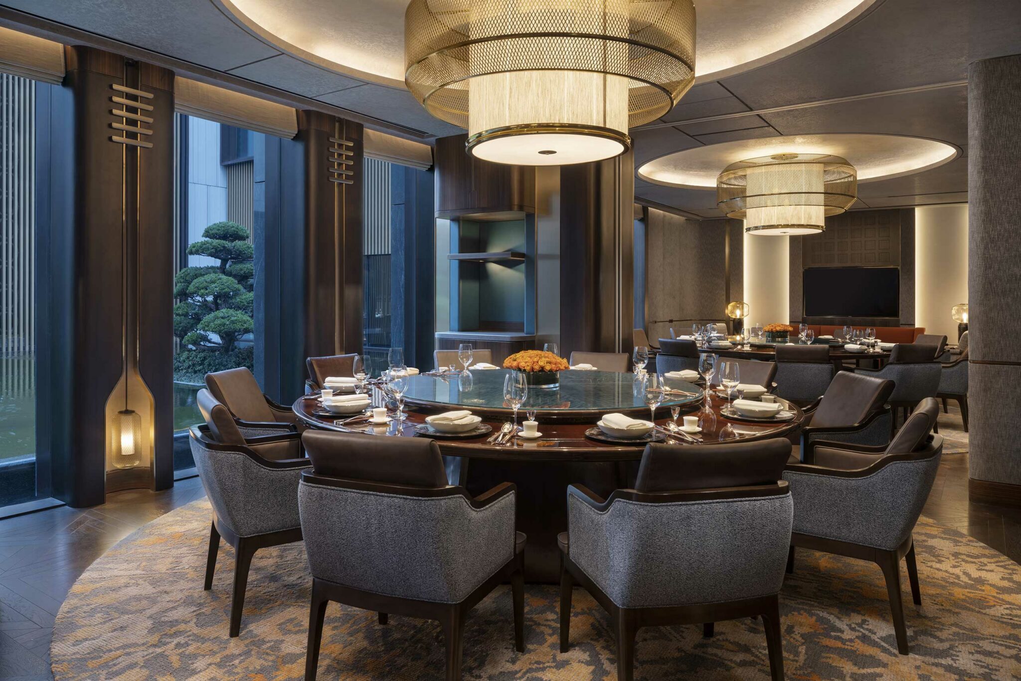 Elegant dining room with a round table set for eight, surrounded by plush chairs. Warm lighting and large windows create a luxurious, welcoming ambiance.