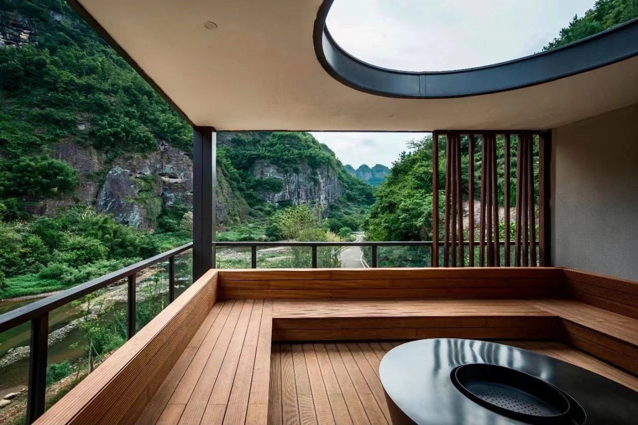 A wooden terrace with a round skylight overlooks lush green mountains and a winding road, conveying a serene and harmonious natural setting.