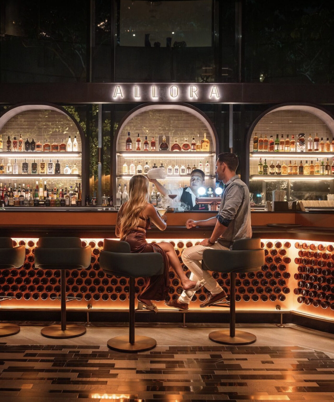 A woman in a red dress and a man in a casual shirt sit at a stylish, well-lit bar named 