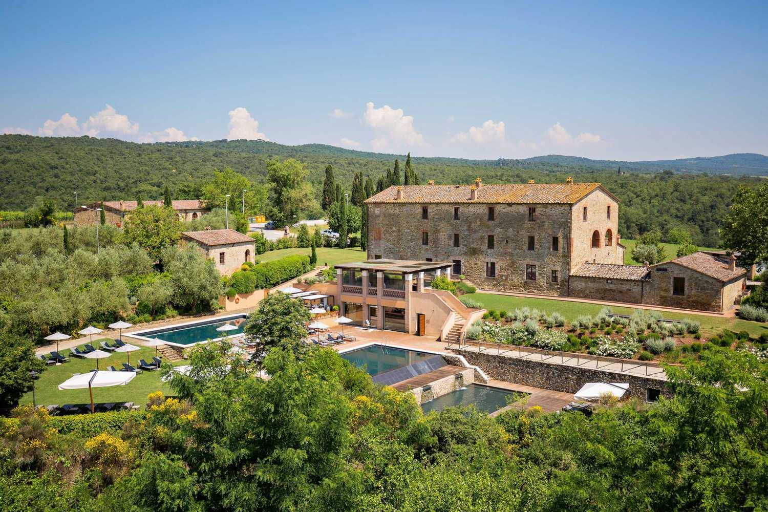 Exterior of Castel Monastero Resort. A rustic villa in Tuscany, Italy, surrounded by lush greenery and hills. It features a large pool, sun loungers, and a relaxing atmosphere under a clear blue sky.