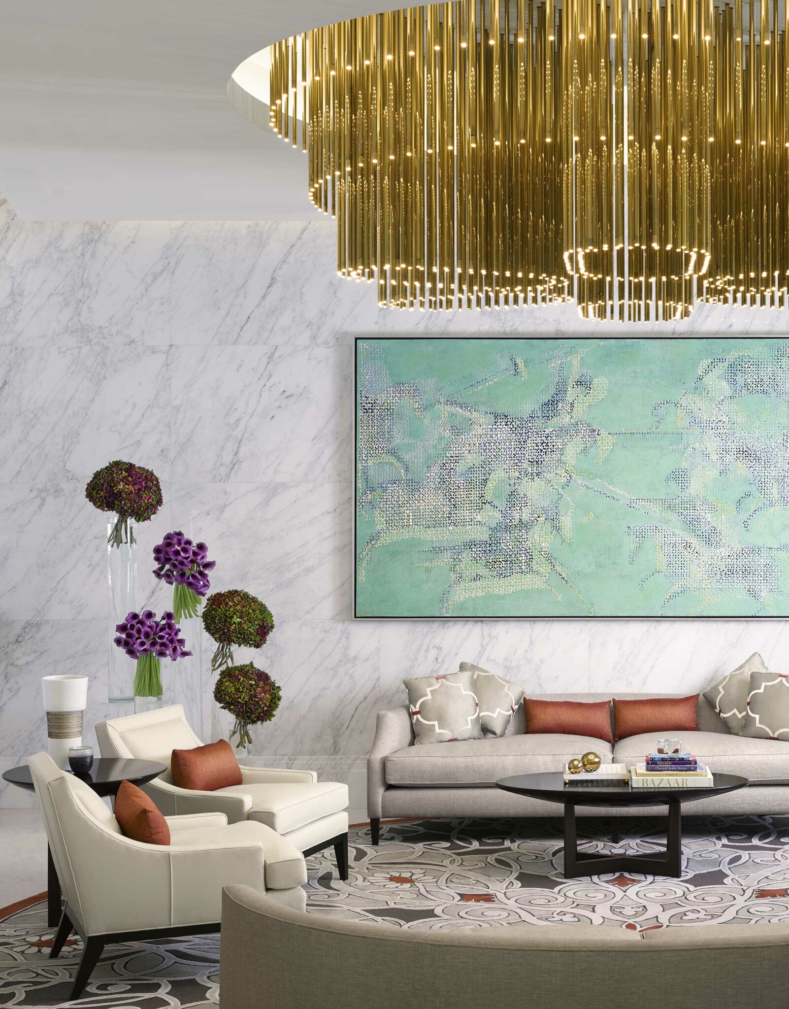 Elegant hotel lobby with a gold chandelier, abstract green art piece, and plush seating. Features marble walls, decorative cushions, and floral displays.