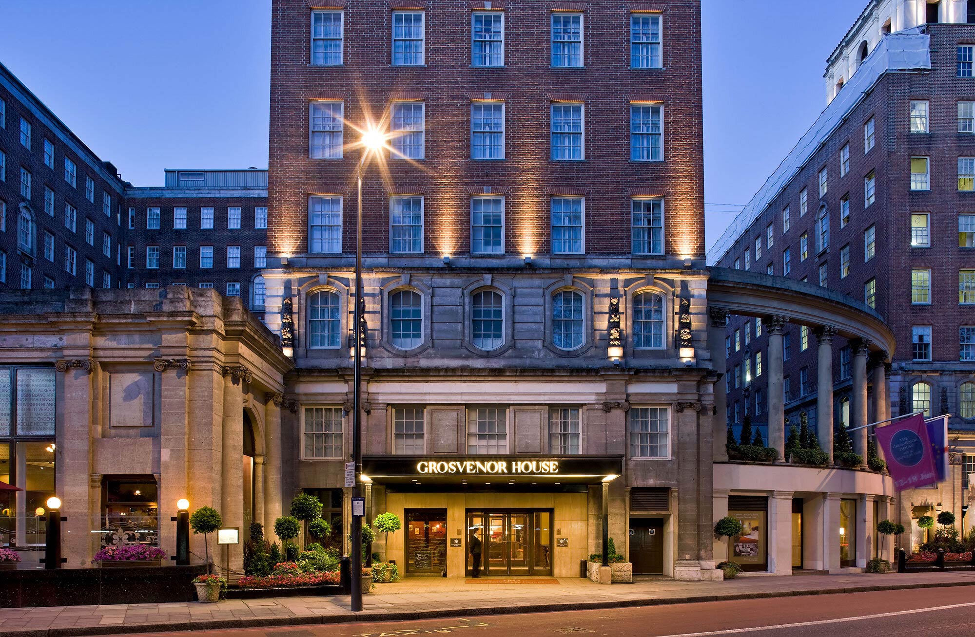 Grand facade of JW Marriott Grosvenor House Hotel, London, at dusk, warmly lit with classic architecture, tall windows, and ornate pillars. Streetlamp glows nearby. Elegant ambiance.