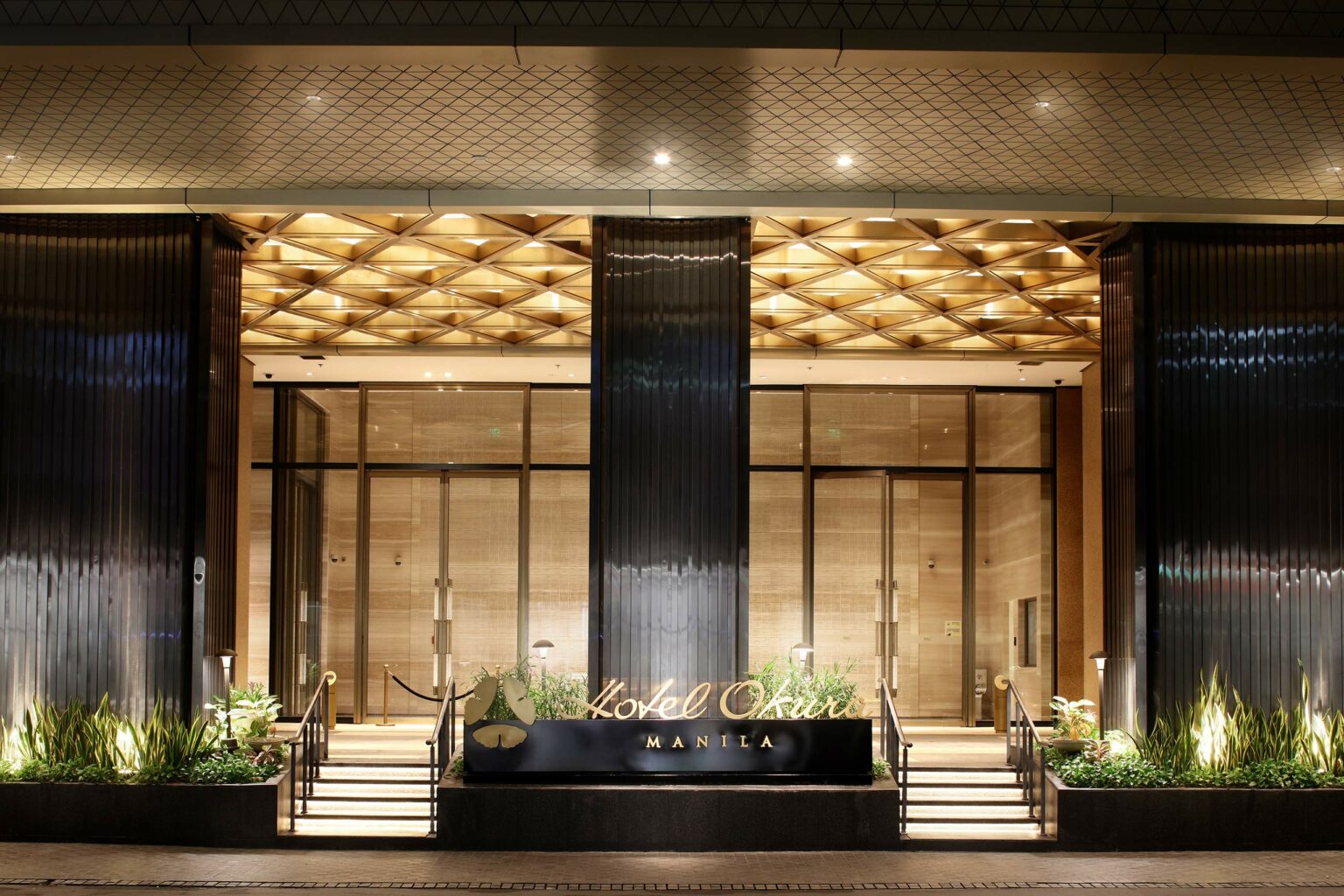 Elegant hotel entrance of Hotel Okura Manila, featuring warm lighting, modern architecture, and a sophisticated atmosphere with a welcoming sign.