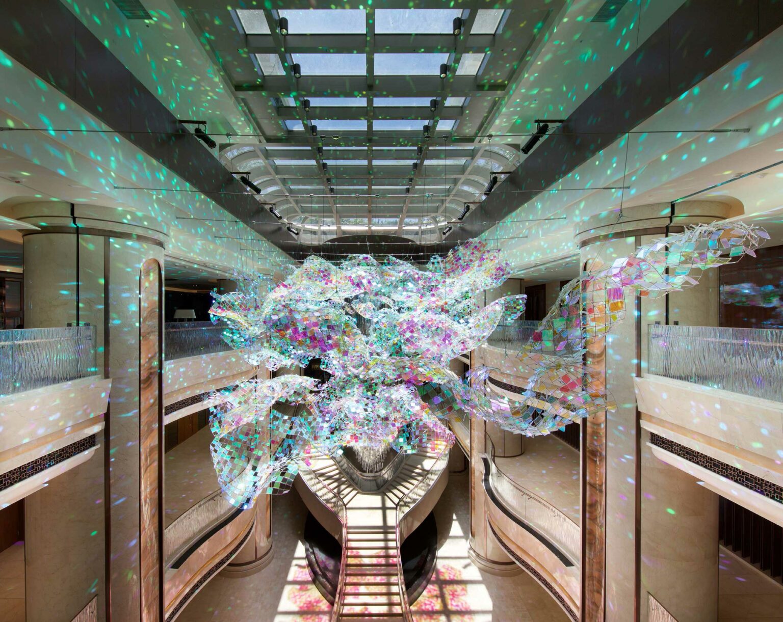 A vibrant, multi-colored chandelier hangs in an elegant hotel atrium. Sunlight filters through, casting dazzling reflections on the beige walls and floor.