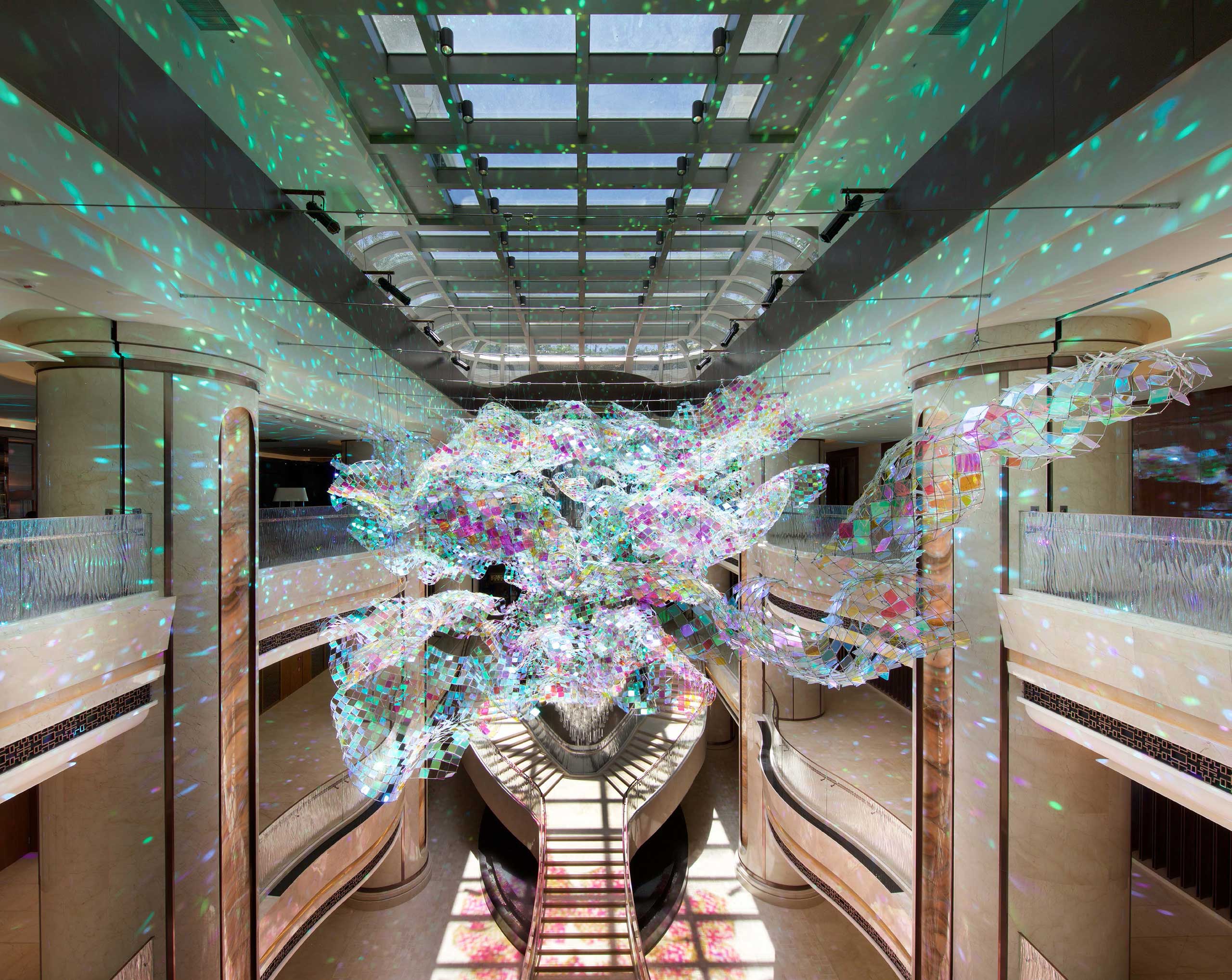 A vibrant, multi-colored chandelier hangs in an elegant hotel atrium. Sunlight filters through, casting dazzling reflections on the beige walls and floor.
