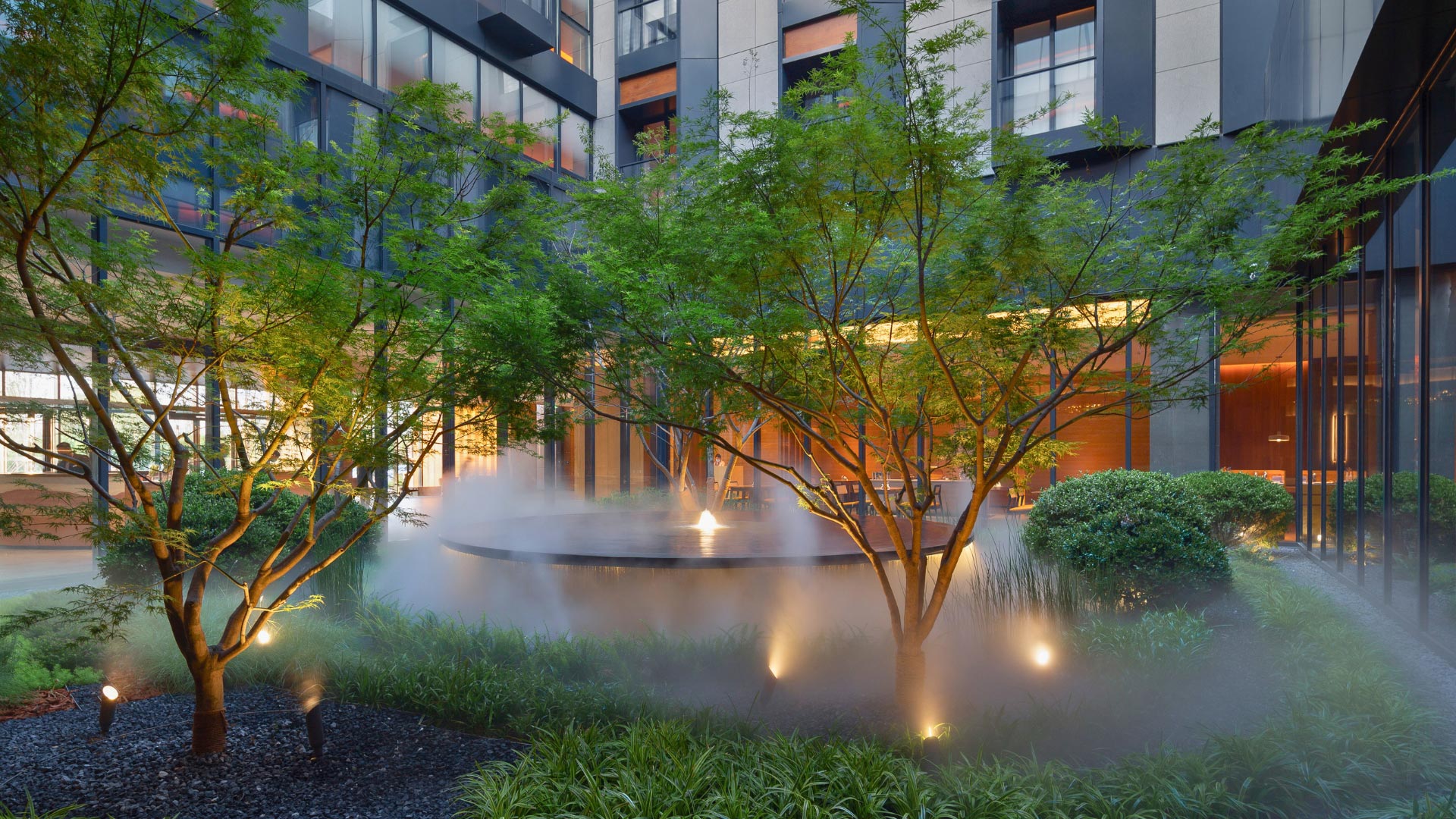 A serene hotel courtyard with lush green trees, soft mist rising, ambient lighting, and a modern building backdrop creates a calm, tranquil atmosphere.