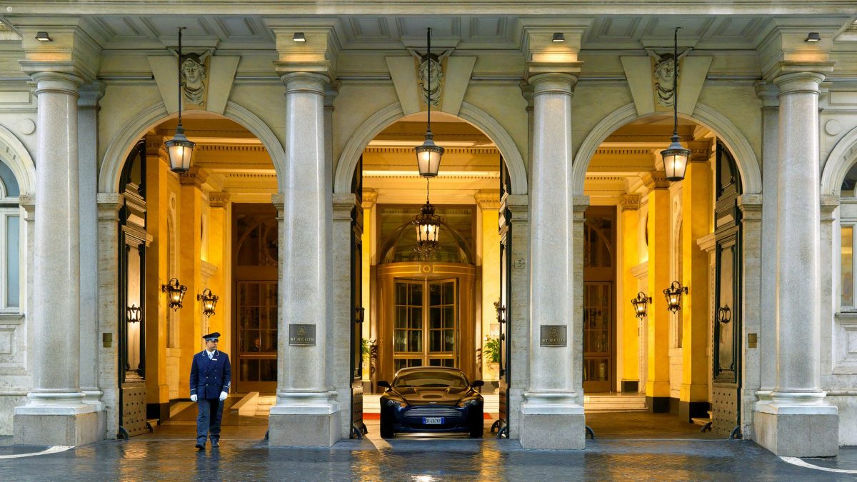 Elegant hotel entrance of The St. Regis Rome, Italy, featuring grand arches and warm lighting. A luxury car is parked centrally, and a doorman in uniform stands to the side, conveying sophistication.
