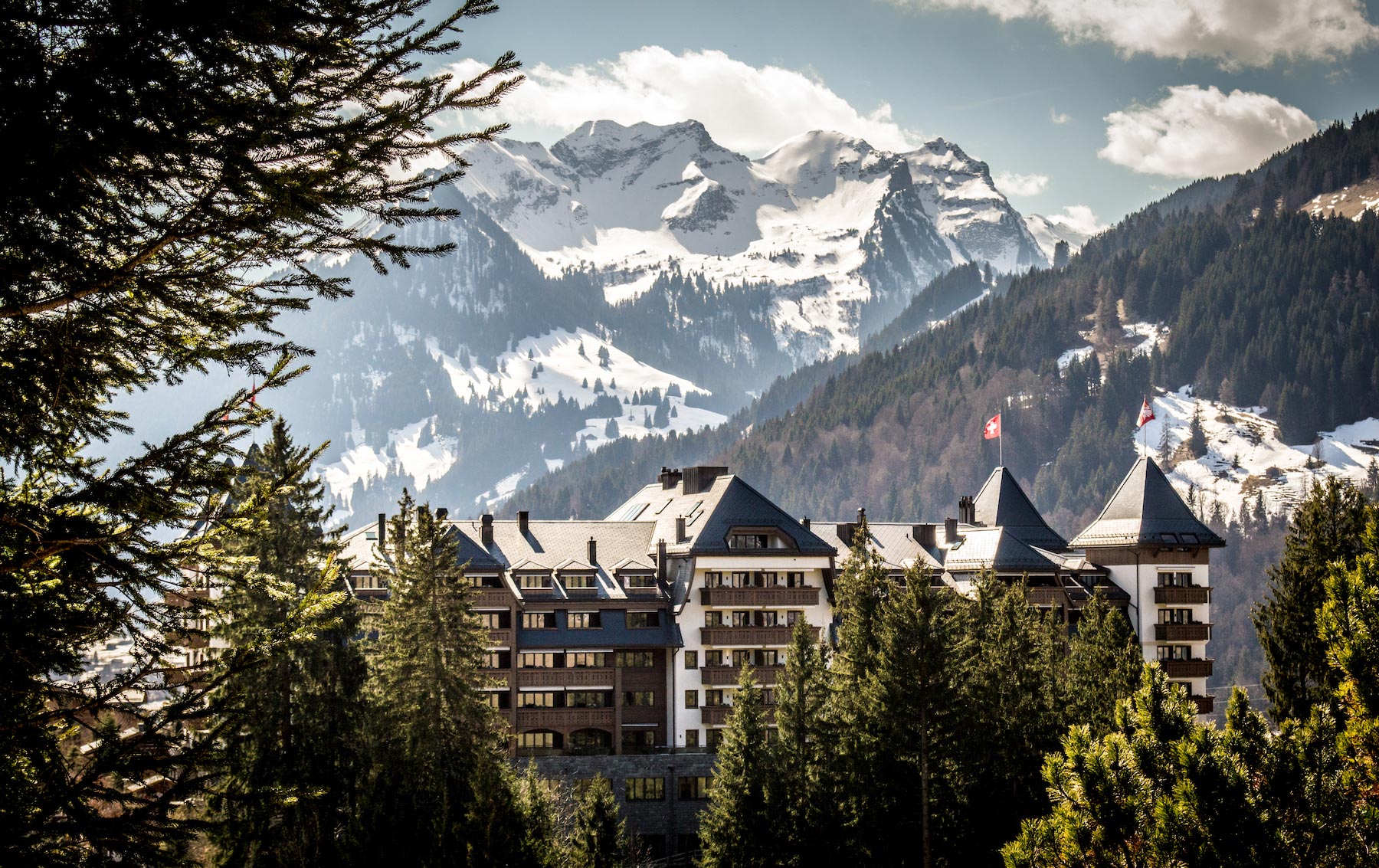 Exterior of The Alpina Gstaad, Switzerland, featuring a majestic mountain hotel surrounded by evergreen trees, set against rugged snow-capped peaks under a partly cloudy sky.