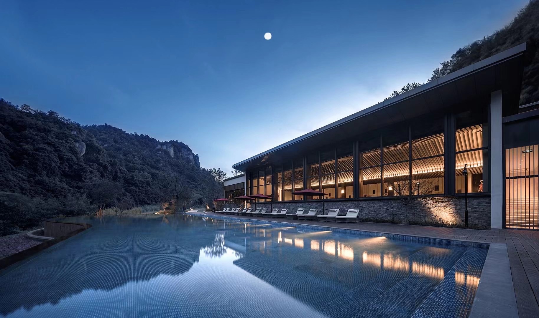 Modern hotel with large windows lit up at dusk, reflecting on a calm infinity pool. Surrounded by hills, with a clear sky and a bright moon above.