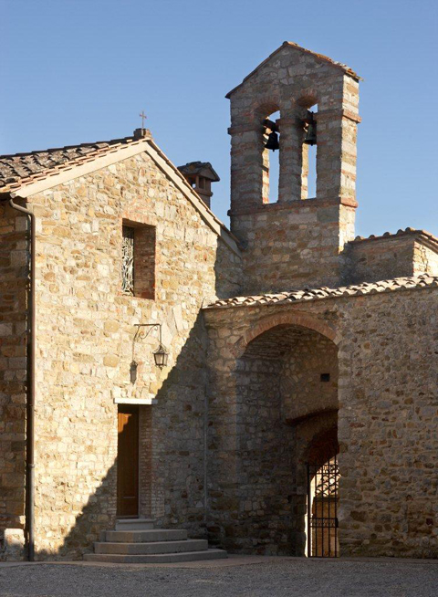 Stone church facade with arched bell tower under clear blue sky. Warm sunlight casts shadows, highlighting rustic texture and weathered charm.