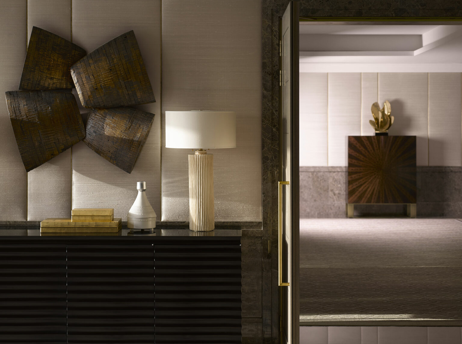 Sophisticated suite entrance featuring a textured wall sculpture, modern lamp, and decorative items on a sleek console. Warm, elegant ambiance.