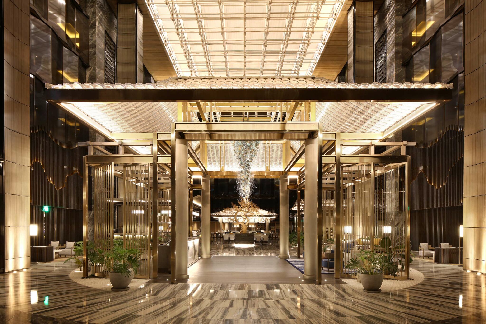 Luxurious hotel lobby featuring a grand golden portal, sculptural chandelier, statement tree installation, and integrated lighting under a vaulted glass ceiling.