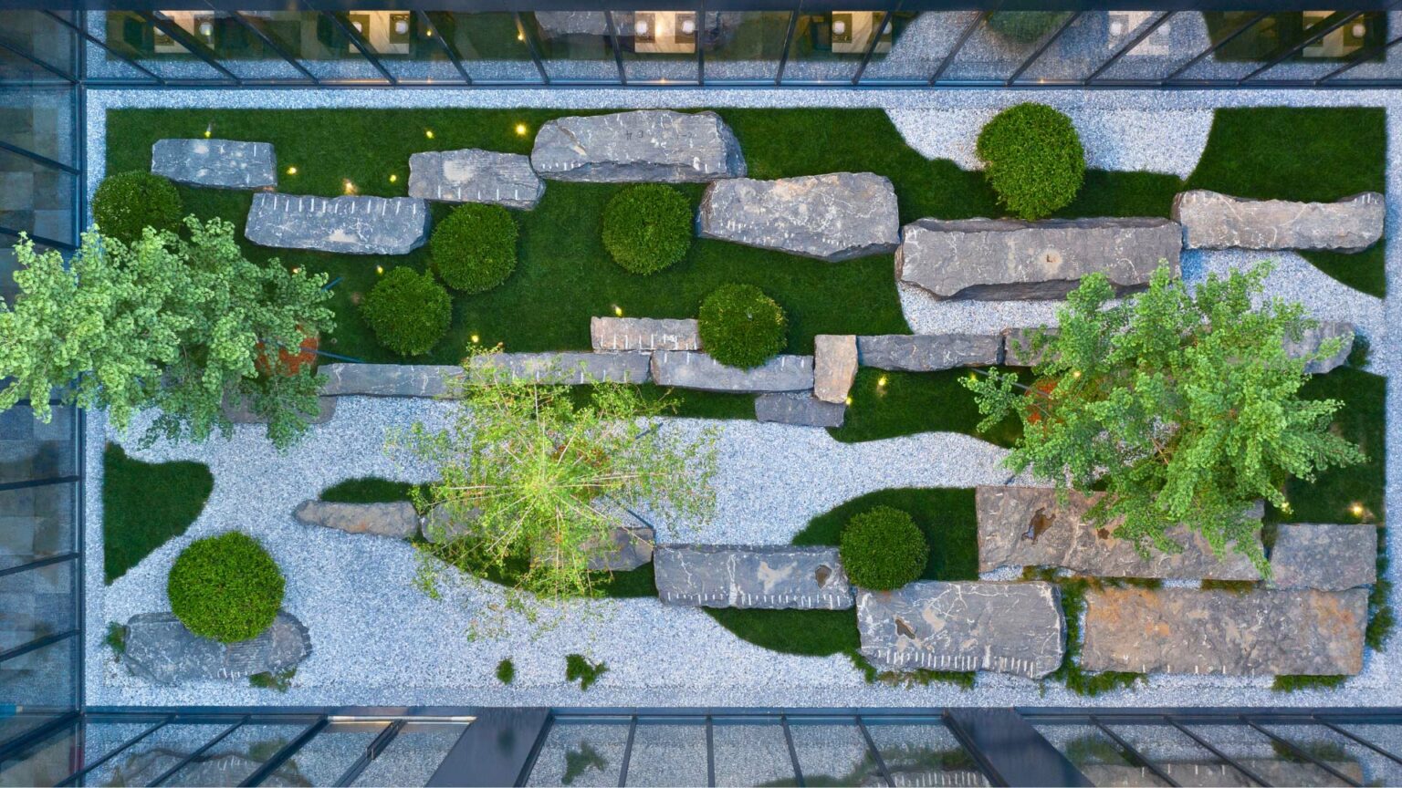 Aerial view of a serene hotel courtyard with lush green trees, round bushes, large grey stones, and white gravel paths, bordered by reflective glass walls.
