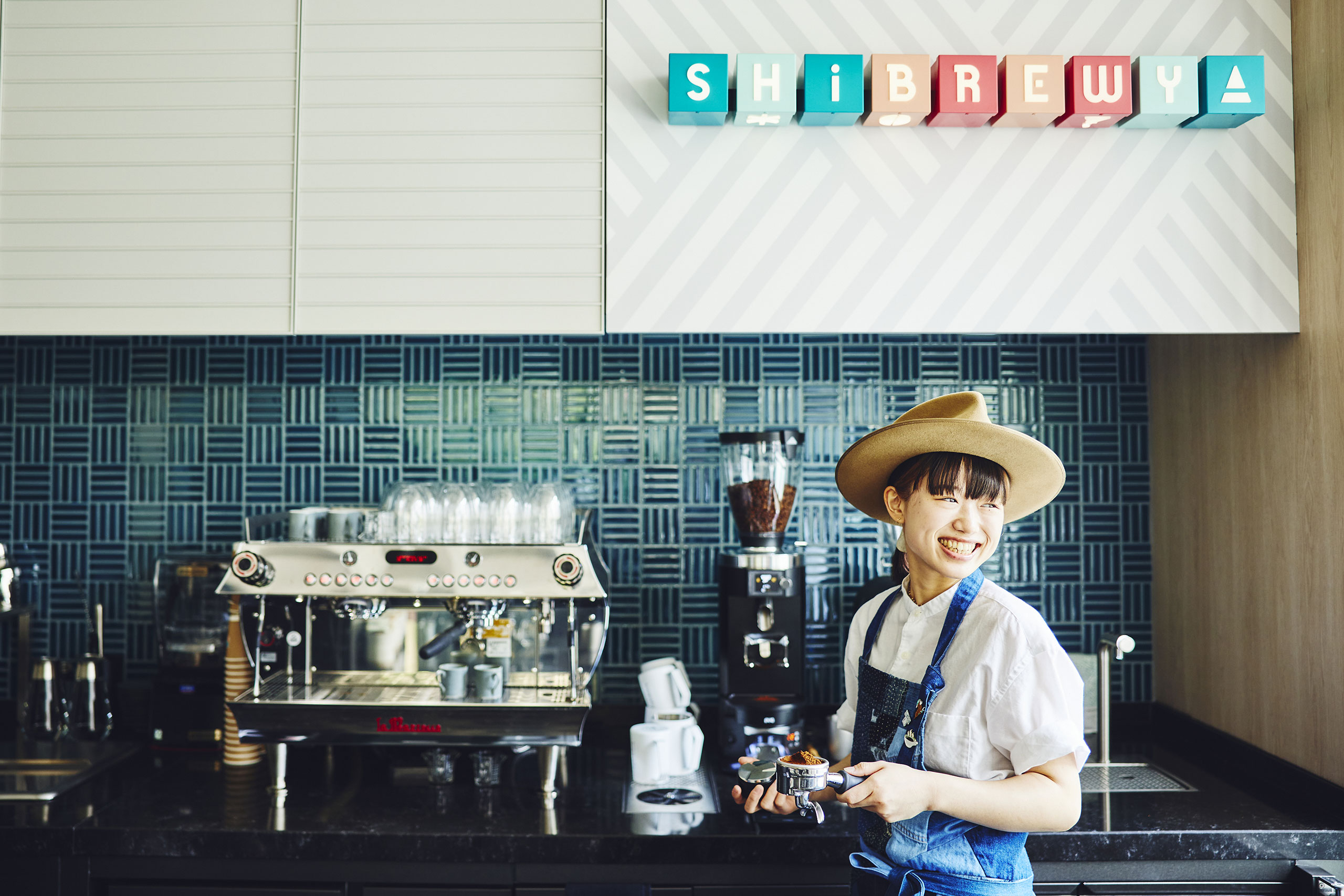 Smiling barista in a hat and apron holds a portafilter in a cheerful cafe. Behind her, espresso machines sit against a stylish blue tile wall. Colorful 3D blocks spelling