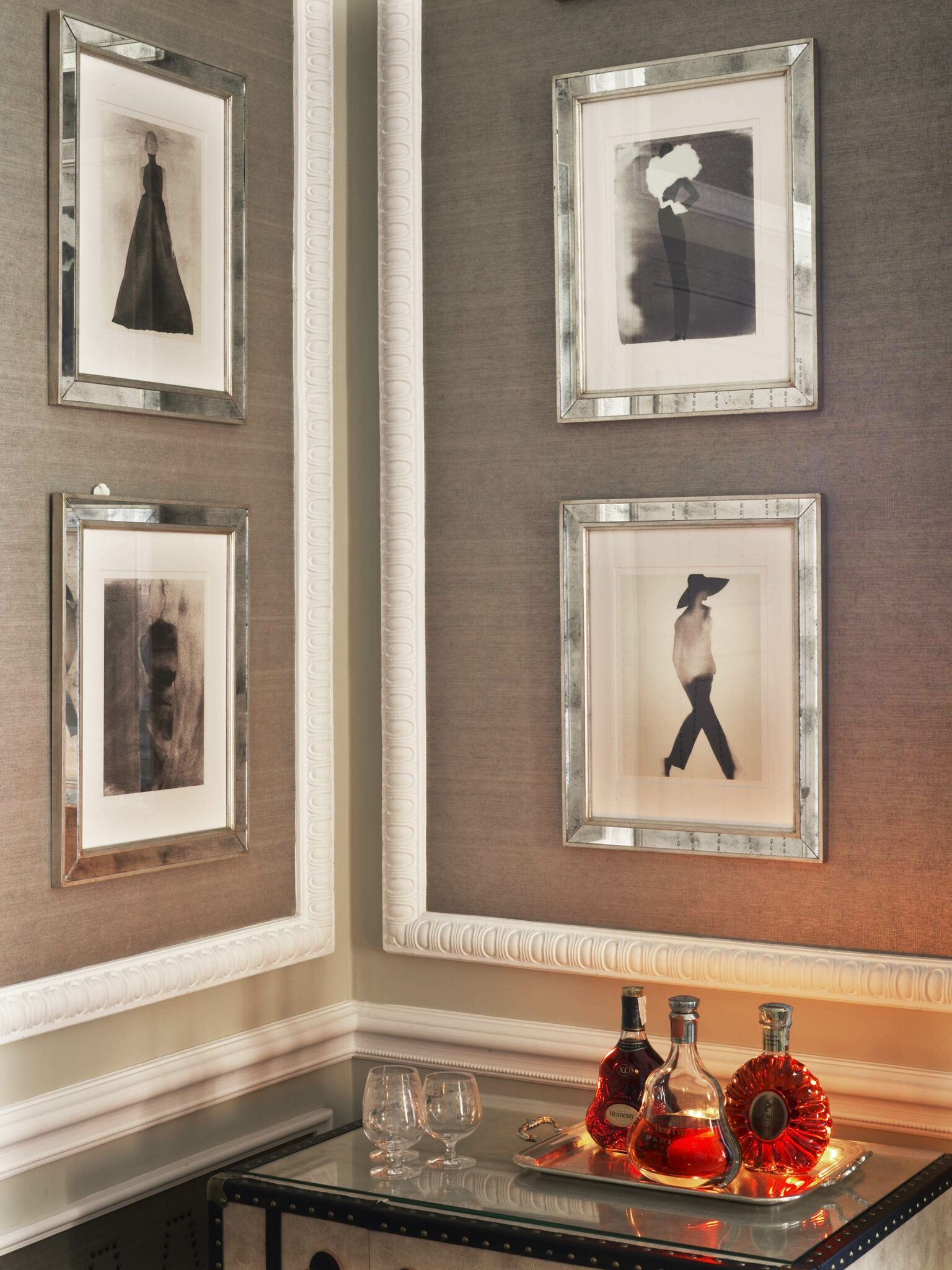 A corner displays four framed black-and-white art pieces on textured walls above a mirrored table. The table holds three elegant decanters, creating a luxurious atmosphere.