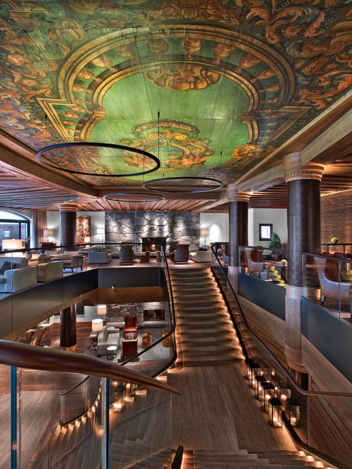 Luxurious hotel lobby with a grand staircase leading to a cozy seating area. A vibrant, ornate ceiling features green and gold patterns, creating an elegant ambiance.