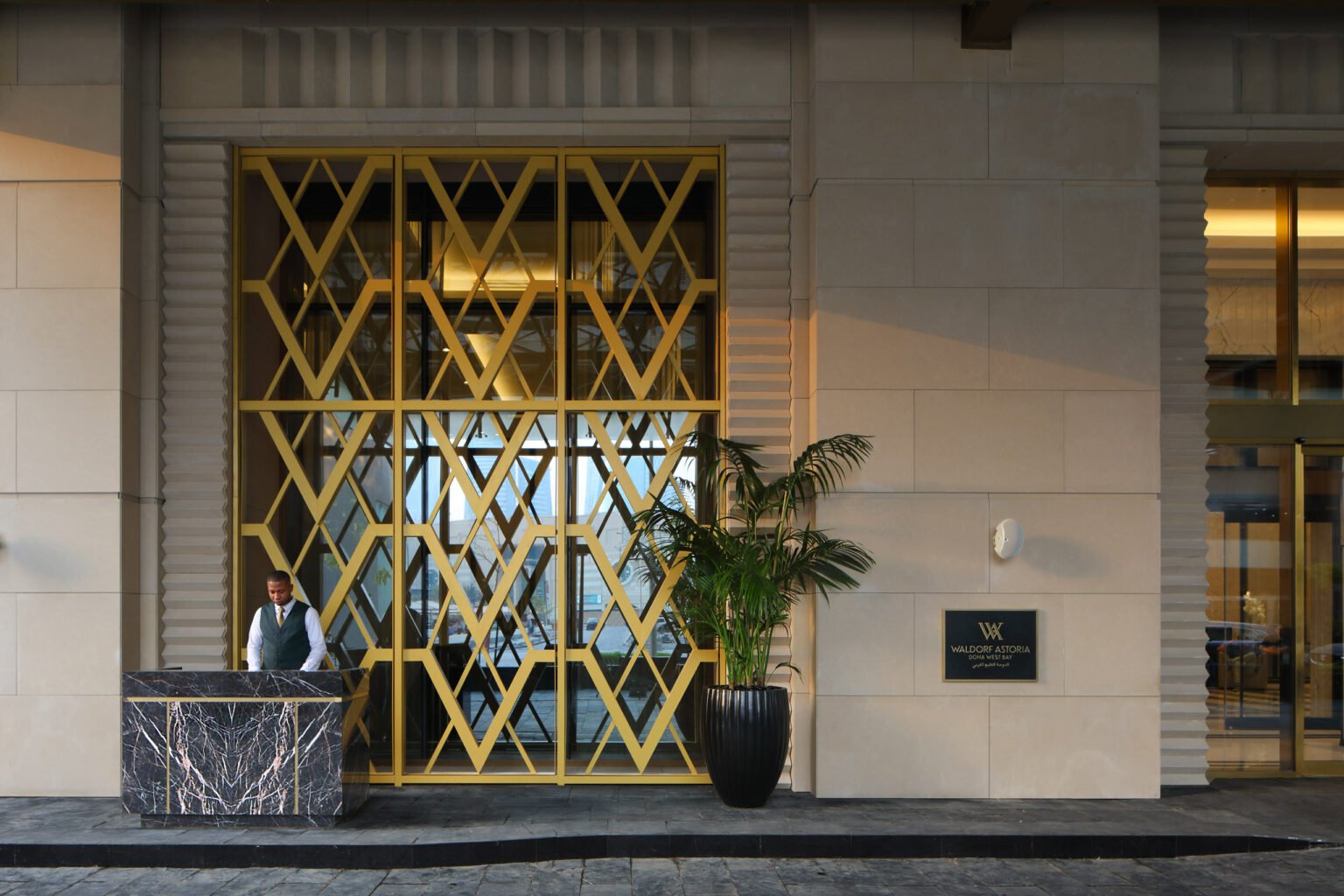 Elegant hotel entrance with gold diamond-patterned doors, a smiling doorman in a green vest by a marble podium, and a large potted plant adding warmth.