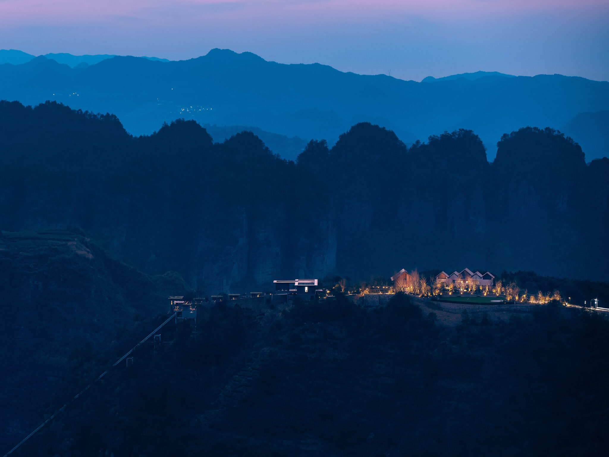 Mountainous landscape at dusk with silhouetted peaks, a softly lit hotel, and glowing trees. A tranquil, serene atmosphere under a pink-tinged sky.