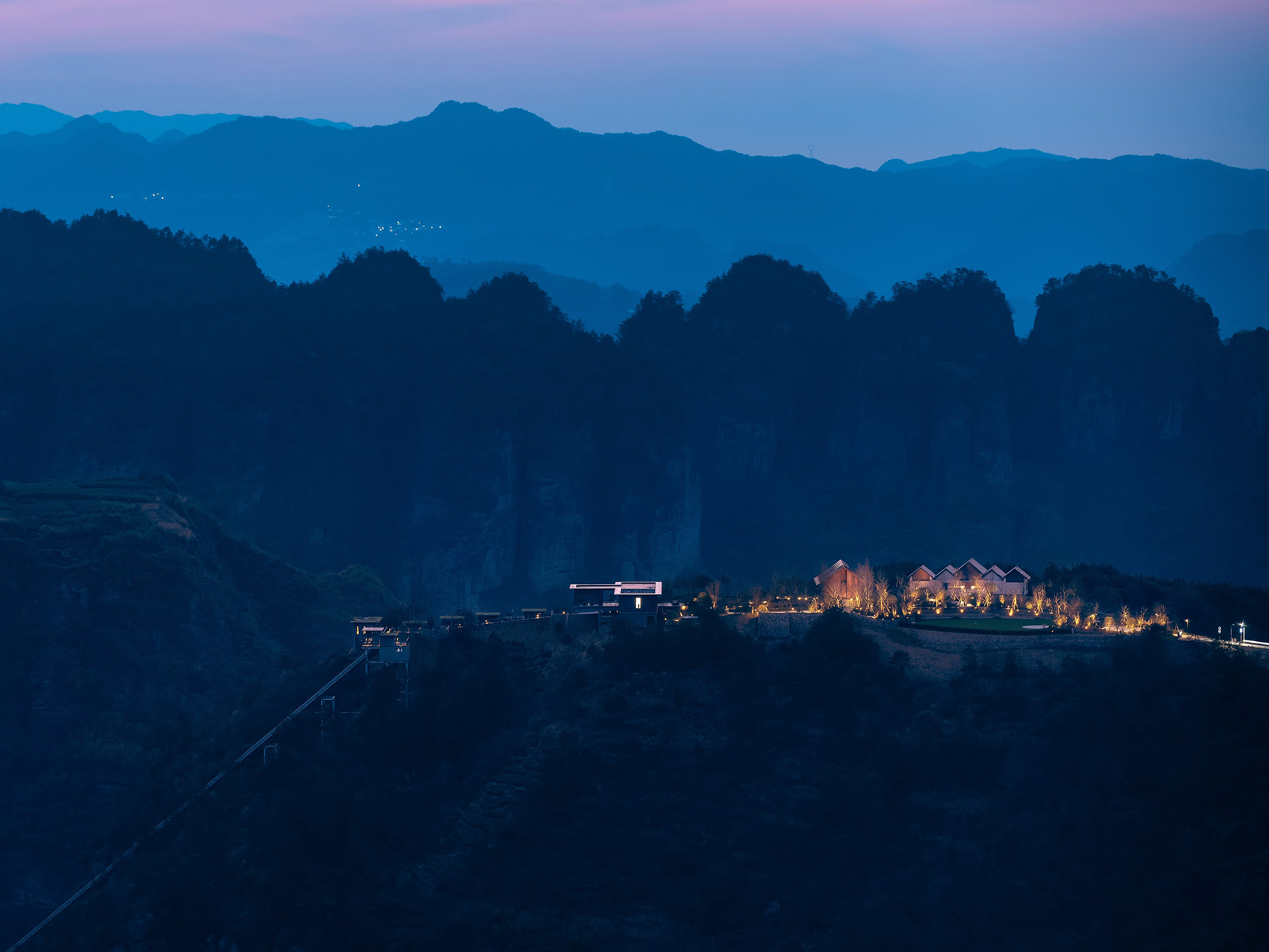 Mountainous landscape at dusk with silhouetted peaks, a softly lit hotel, and glowing trees. A tranquil, serene atmosphere under a pink-tinged sky.