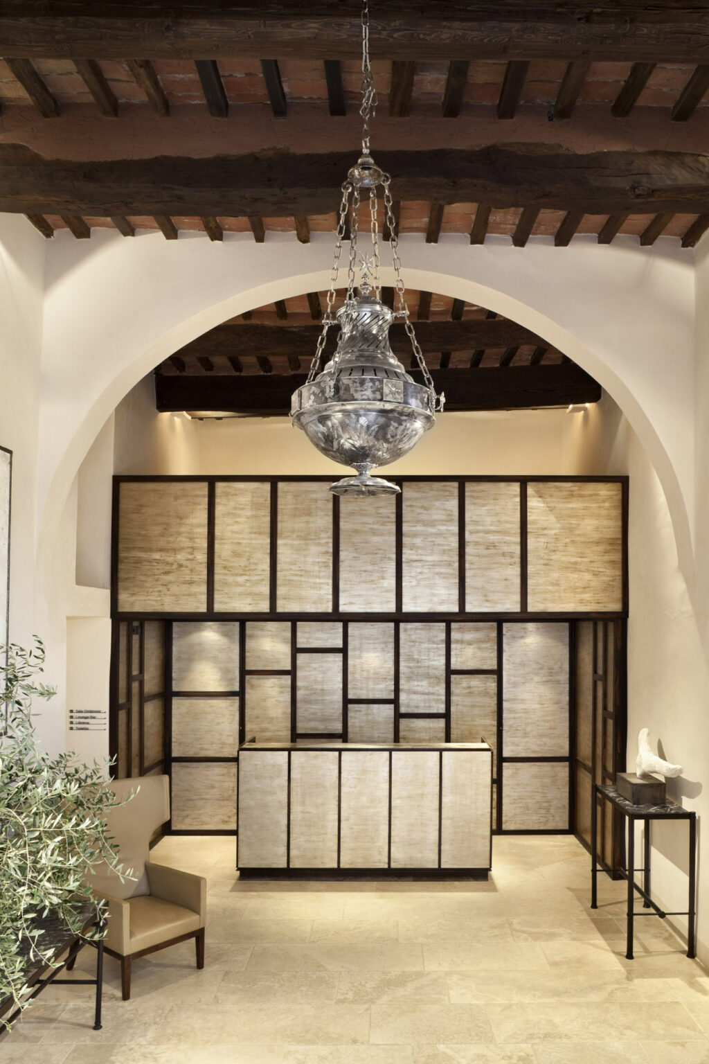 Elegant hotel reception area with a rustic wooden ceiling, large metallic chandelier, beige paneled walls, and minimalist decor, conveying a serene ambiance.
