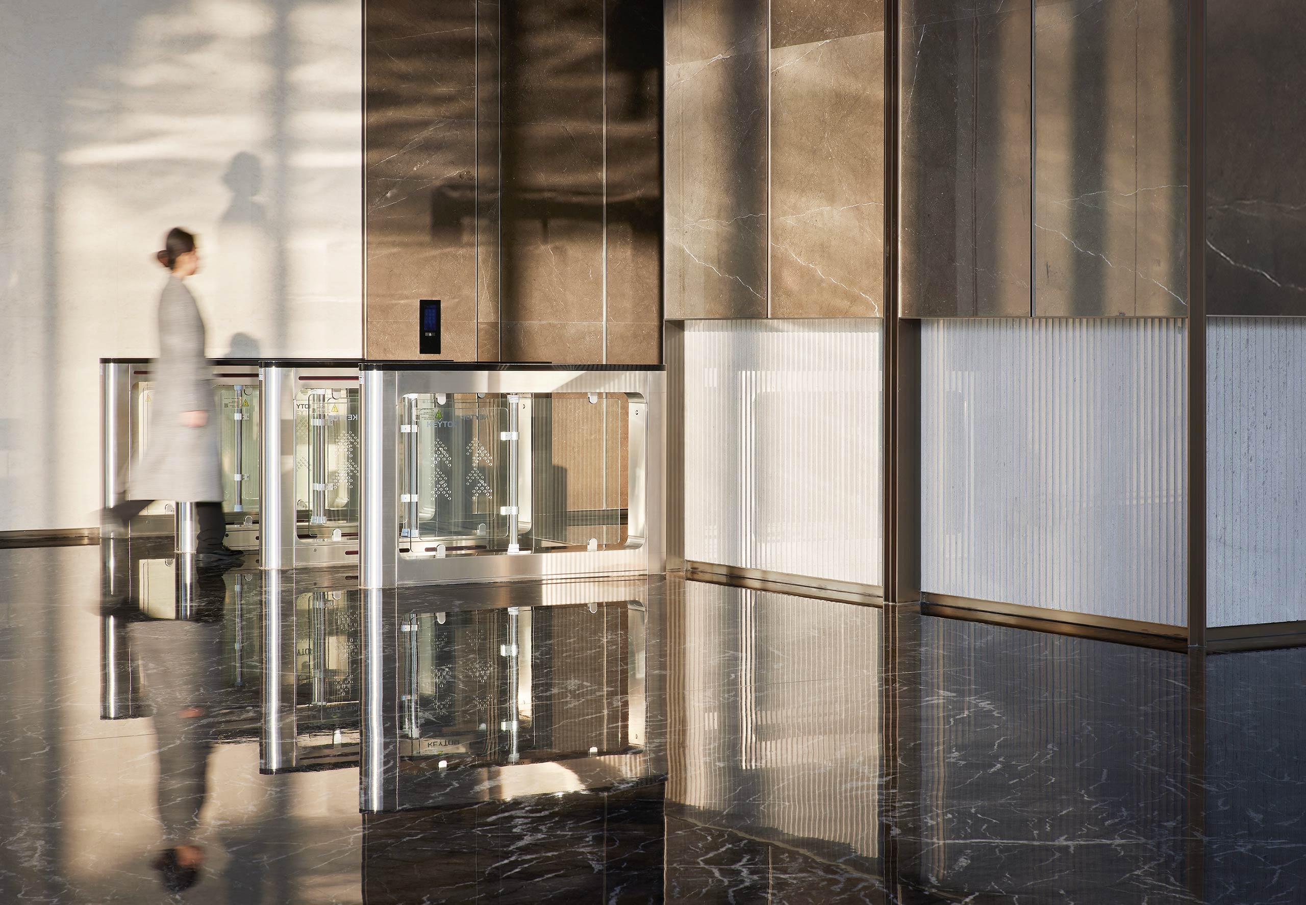 A person in a gray coat walks through a modern office lobby with glossy marble floors and sleek barriers. Soft natural light casts shadows, creating an elegant atmosphere.