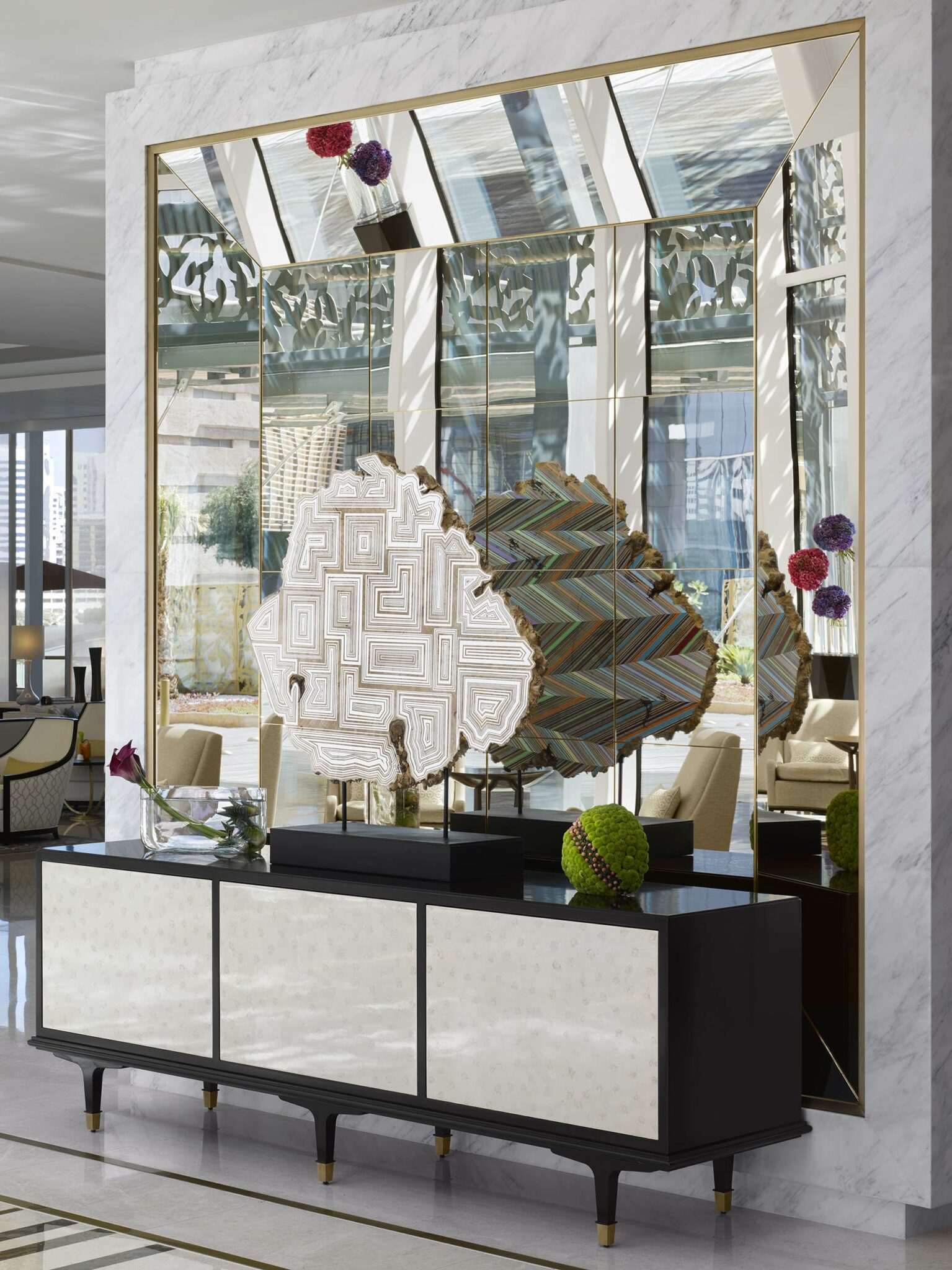 Modern hotel corridor close-up with a mirrored wall reflecting a geometric sculpture and colorful vase. Elegant black and white sideboard below, creating a sleek, artistic ambiance.