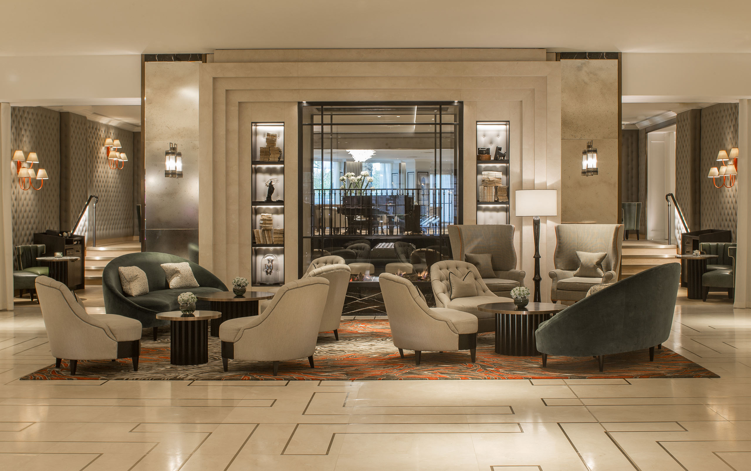 Elegant hotel lobby with plush gray seating, a central coffee table, soft lighting, and decorative shelves. The atmosphere is warm and inviting.