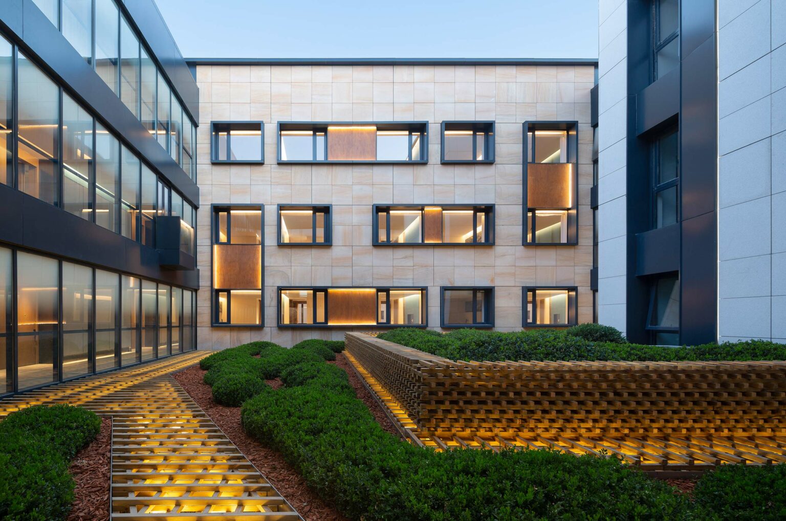 Modern hotel courtyard with sleek buildings framing a garden. Geometric windows and warm lighting create a welcoming, contemporary ambiance.