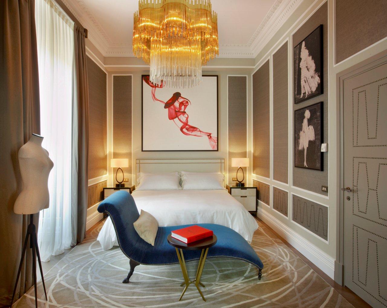 Elegant suite bedroom with a luxurious chandelier, minimalistic decor. Features a navy chaise lounge and beige pillow, beside an art deco gold side table with a red book and a mannequin. Sophisticated and cozy ambiance.