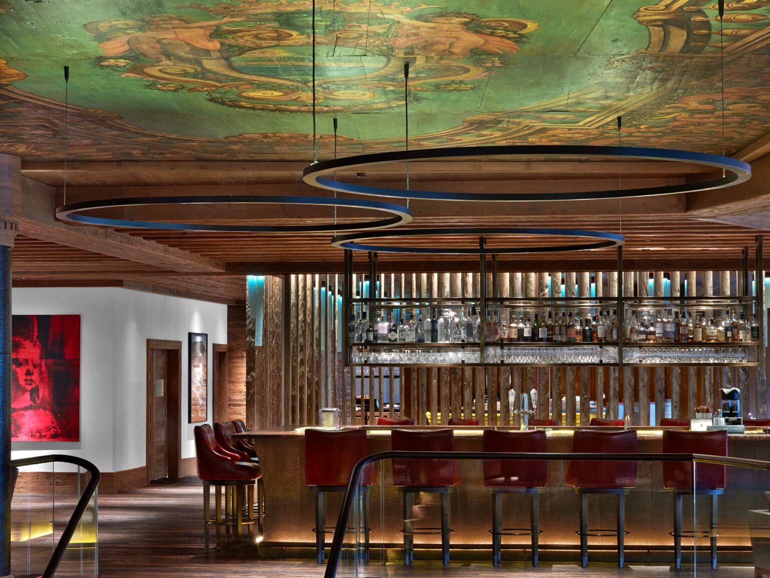 Luxurious hotel bar interior with circular ceiling lights and mural. Features a stocked bar, red barstools, and warm wood accents, creating an upscale ambiance.