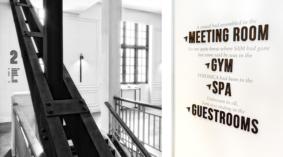 Black and white hotel interior showing a staircase with bold lettering on the wall pointing to rooms like 