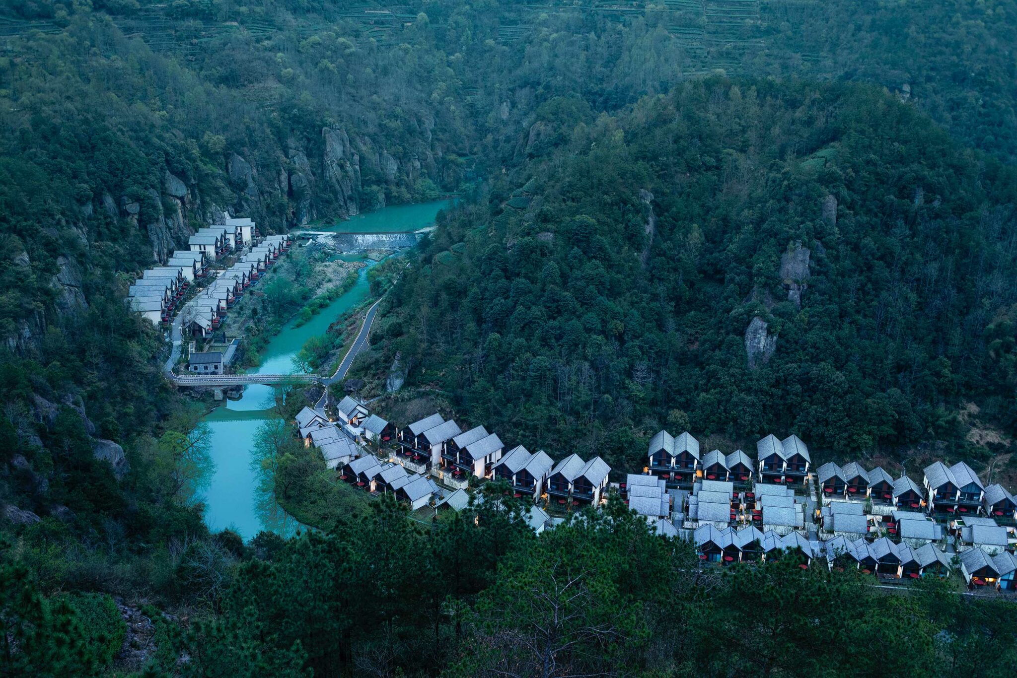 Aerial view of a serene village nestled in lush green hills, with winding turquoise river and neat rows of white-roofed houses, evoking tranquility.